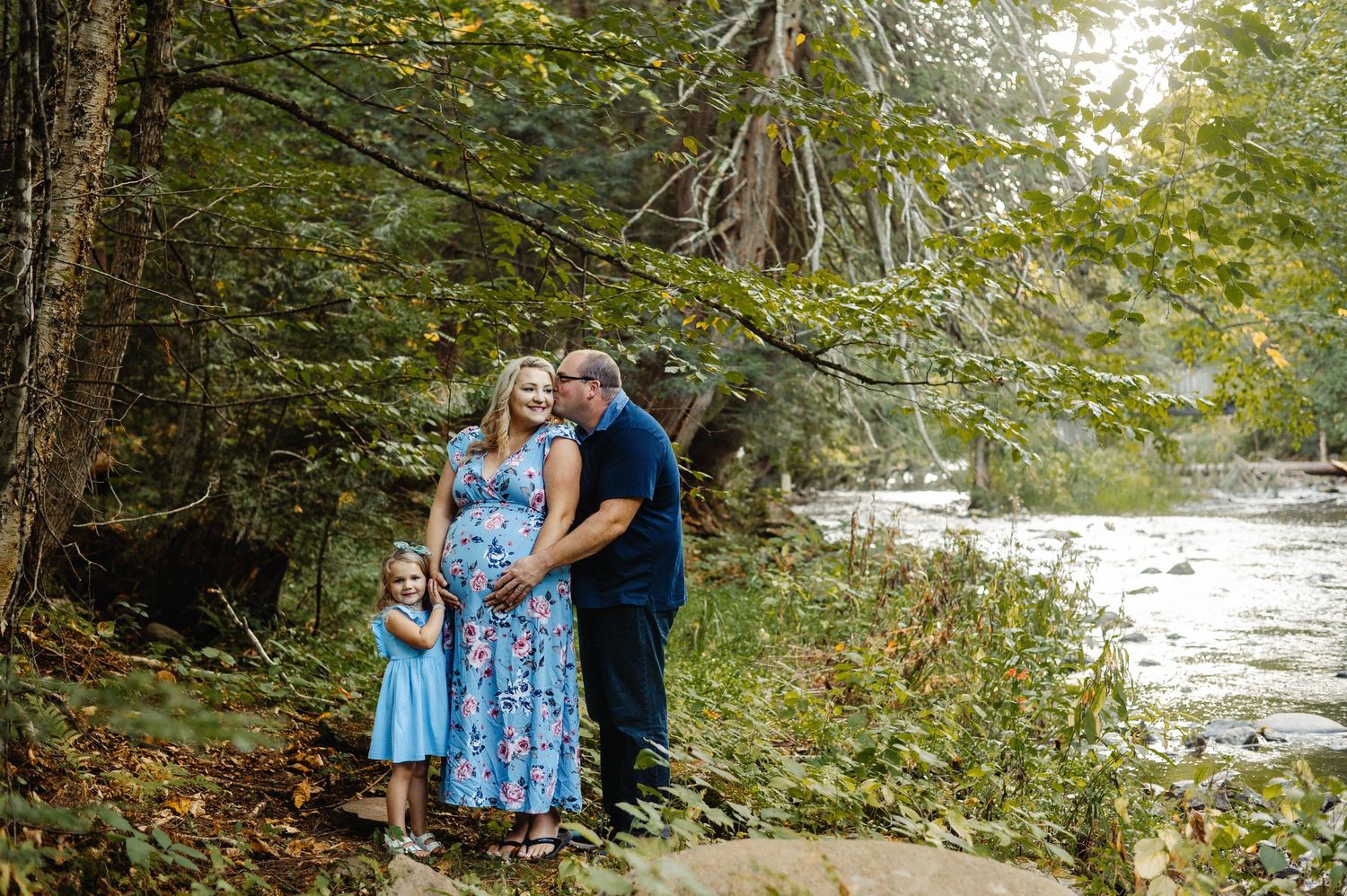 Family cuddled by the waters edge on the river while mom holds baby belly. Ottawa Maternity Photographer. Ottawa Family Photography