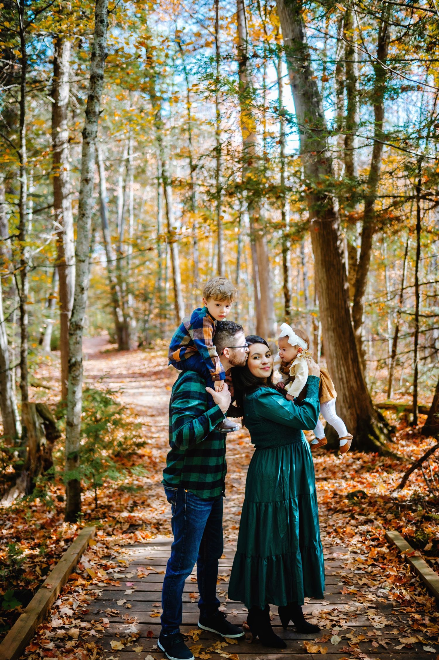 Son on dad's shoulders while he kisses mom while she holds daughter in the fall leaves. Ottawa Family Photography.