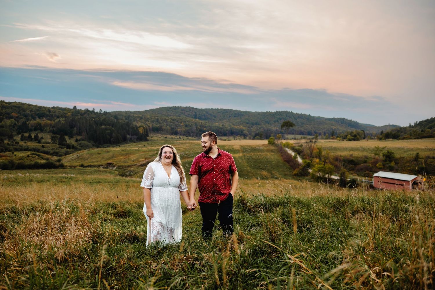 Engaged couple walking in a field with covered bridge in background. Ottawa Wedding Photographer. Ottawa Couples Photographer