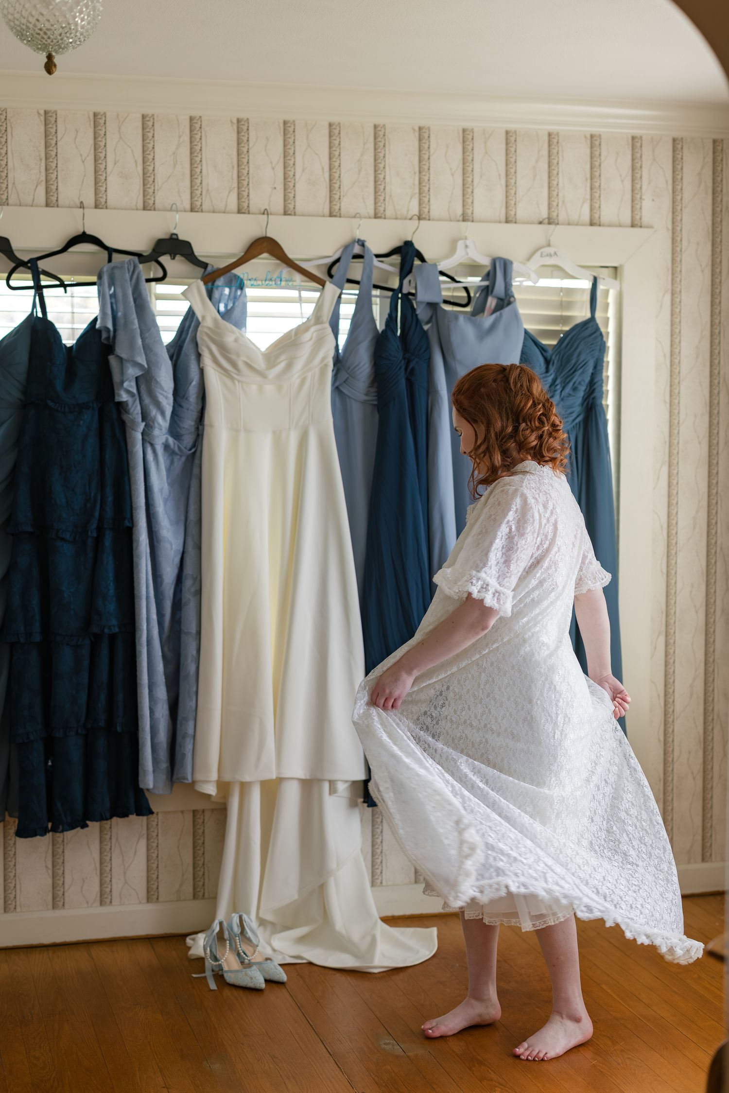 Bride in white lace robe twirling before her wedding gown, surrounded by blue bridesmaid dresses hanging on a wall.