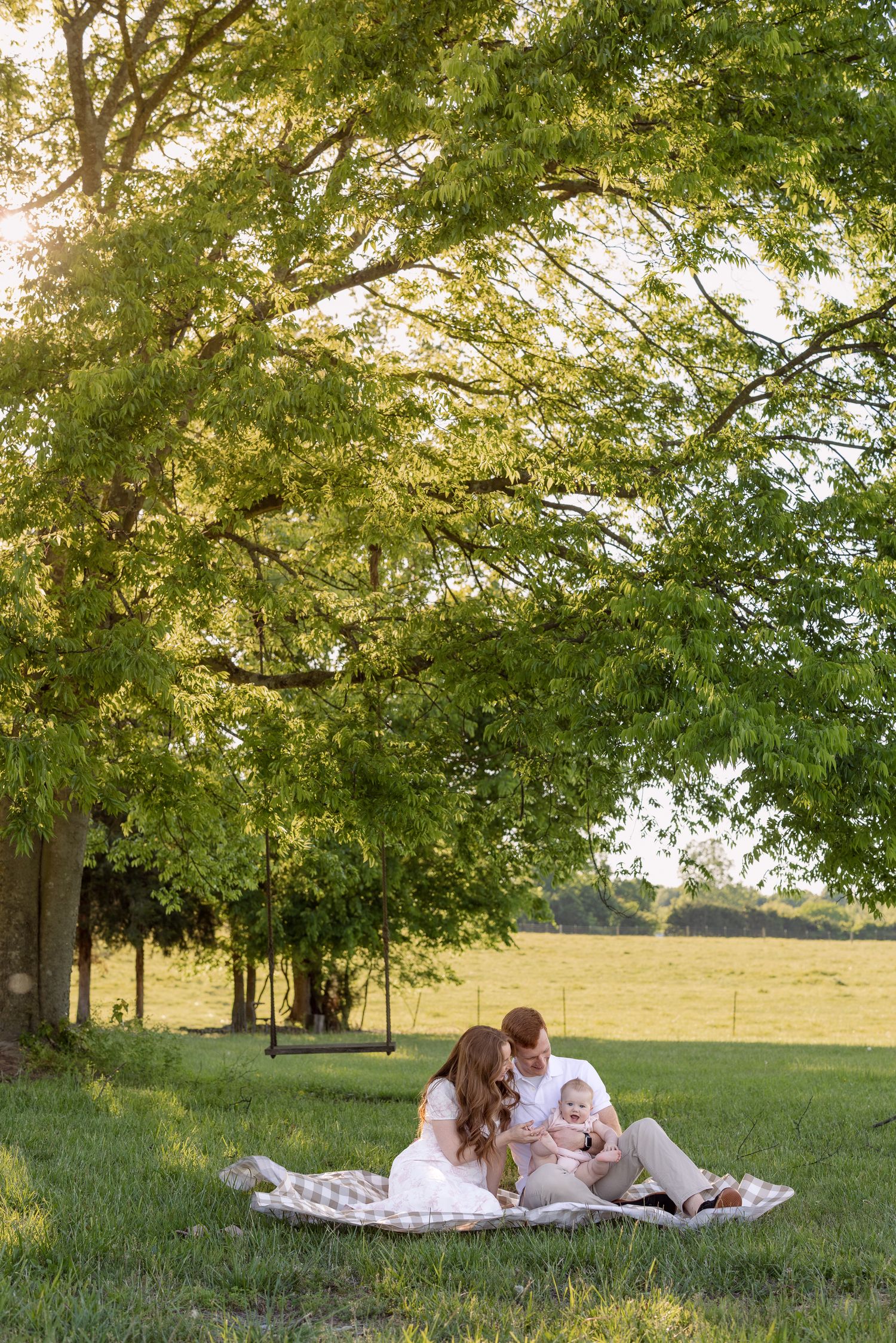 Young family sitting on a blanket under a large green tree in a sunny field, parents smiling at their baby.