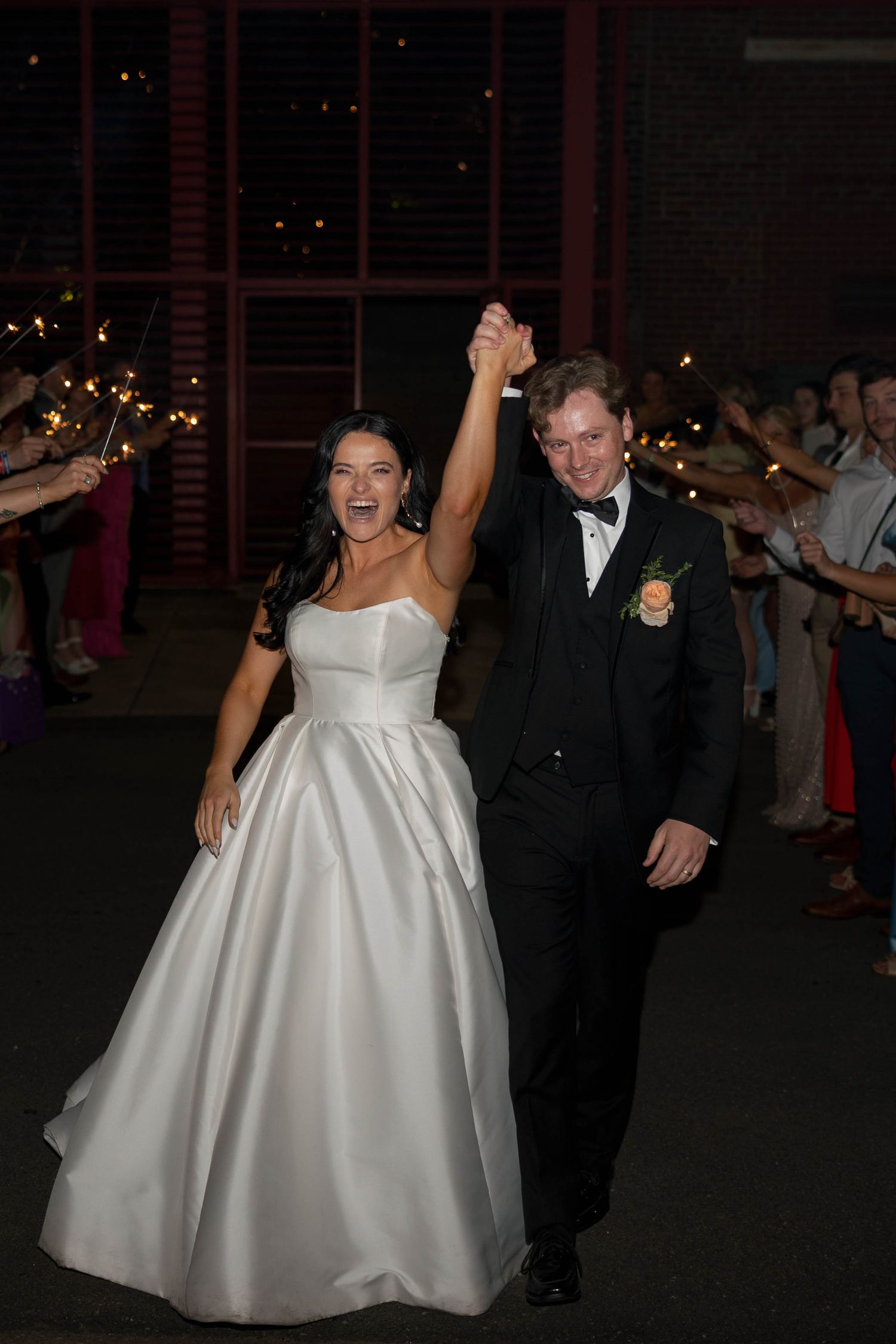 Joyful newlyweds exit wedding reception through sparkler tunnel, bride in white ballgown and groom in black tuxedo raising hands.