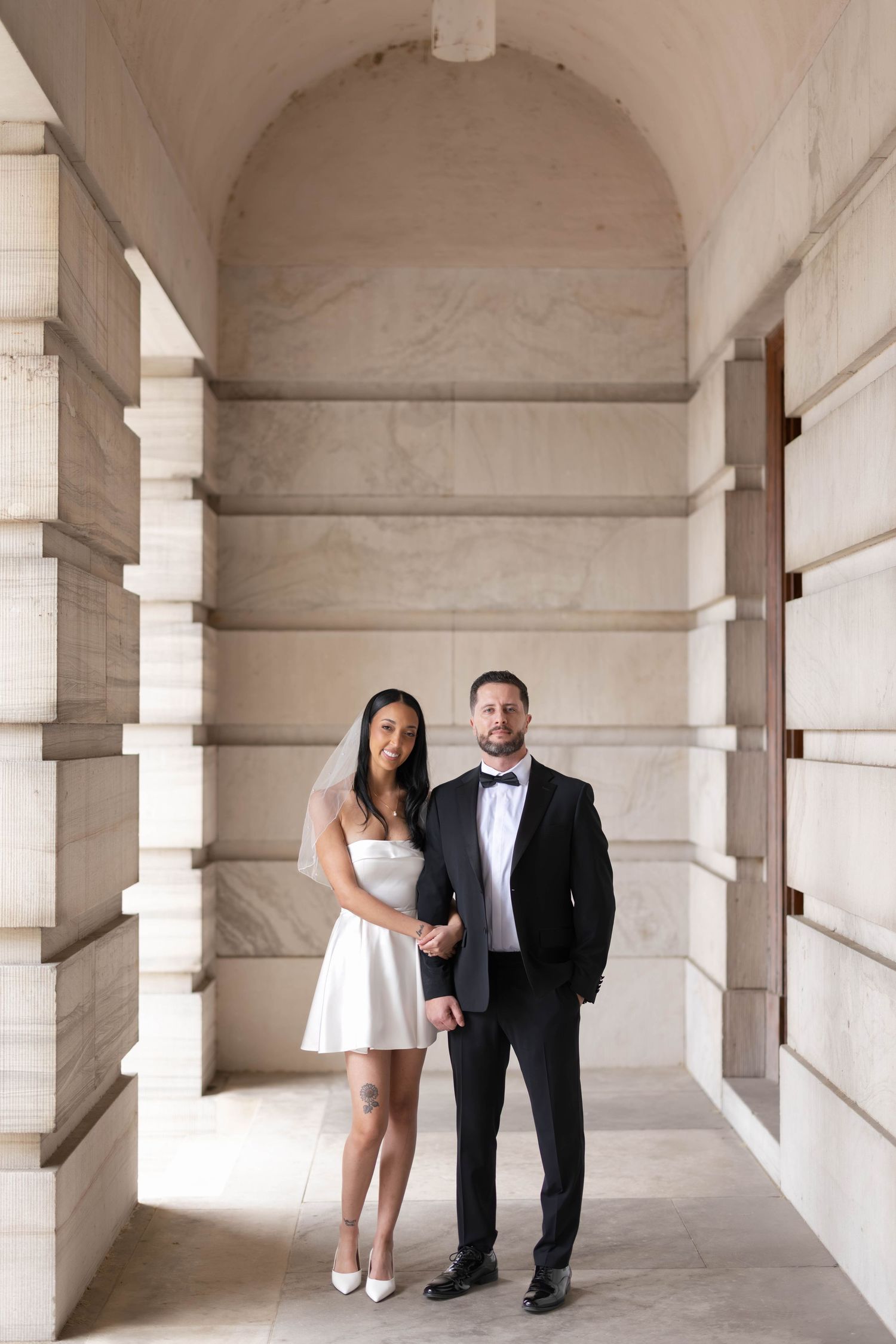 Bride in short white dress and groom in black tuxedo pose in elegant marble hallway for wedding portrait.