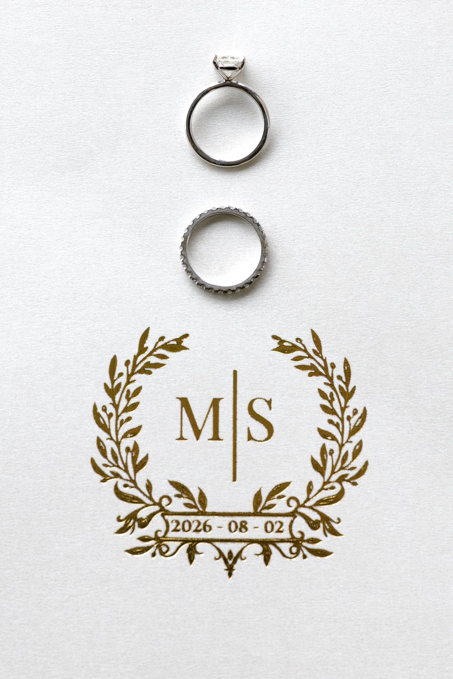 Two silver wedding rings beside a gold monogram crest with initials M|S and wedding date 2026-08-02 on white background.