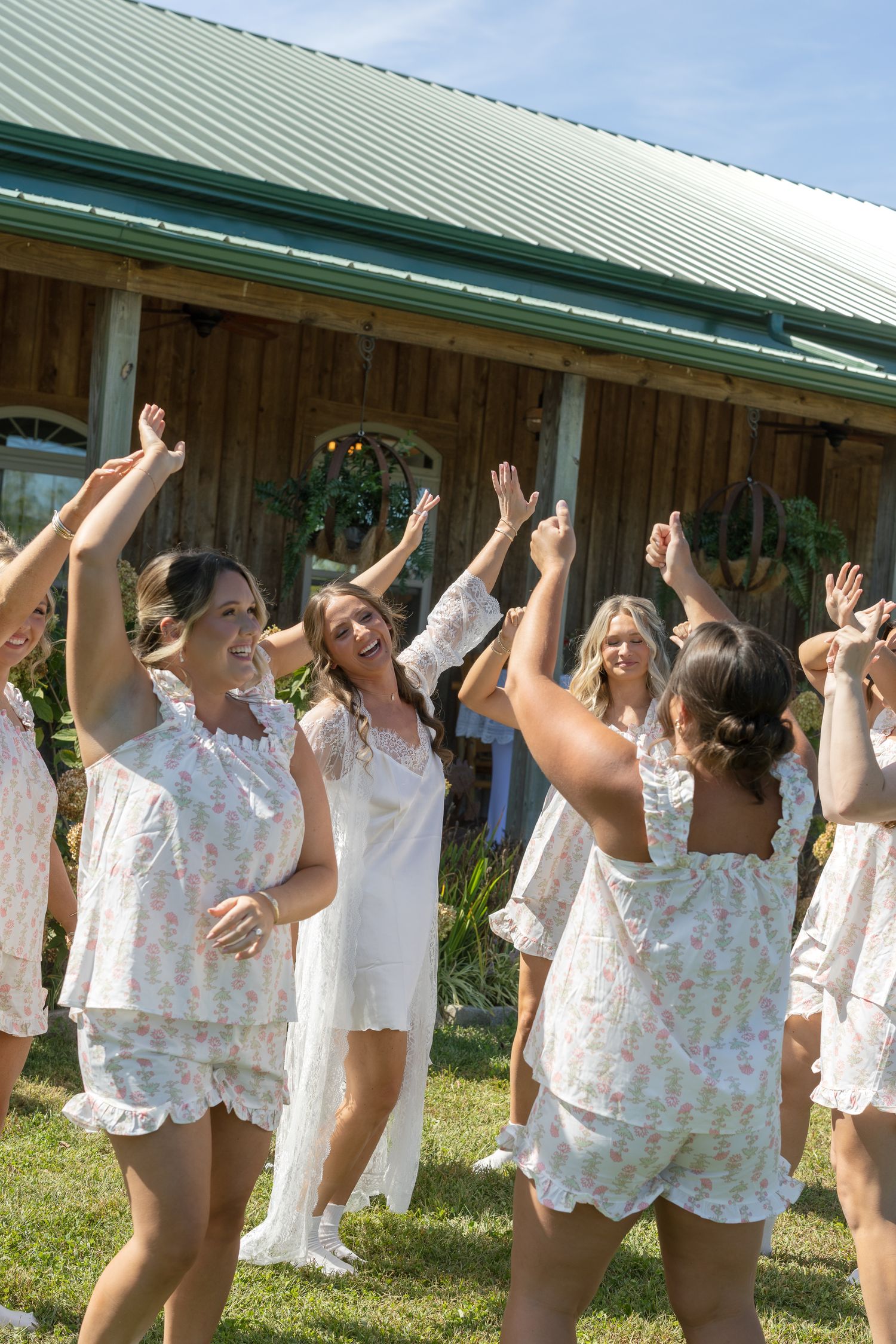 Bride in white gown celebrates with bridesmaids in floral pajamas, arms raised joyfully outside rustic barn venue.