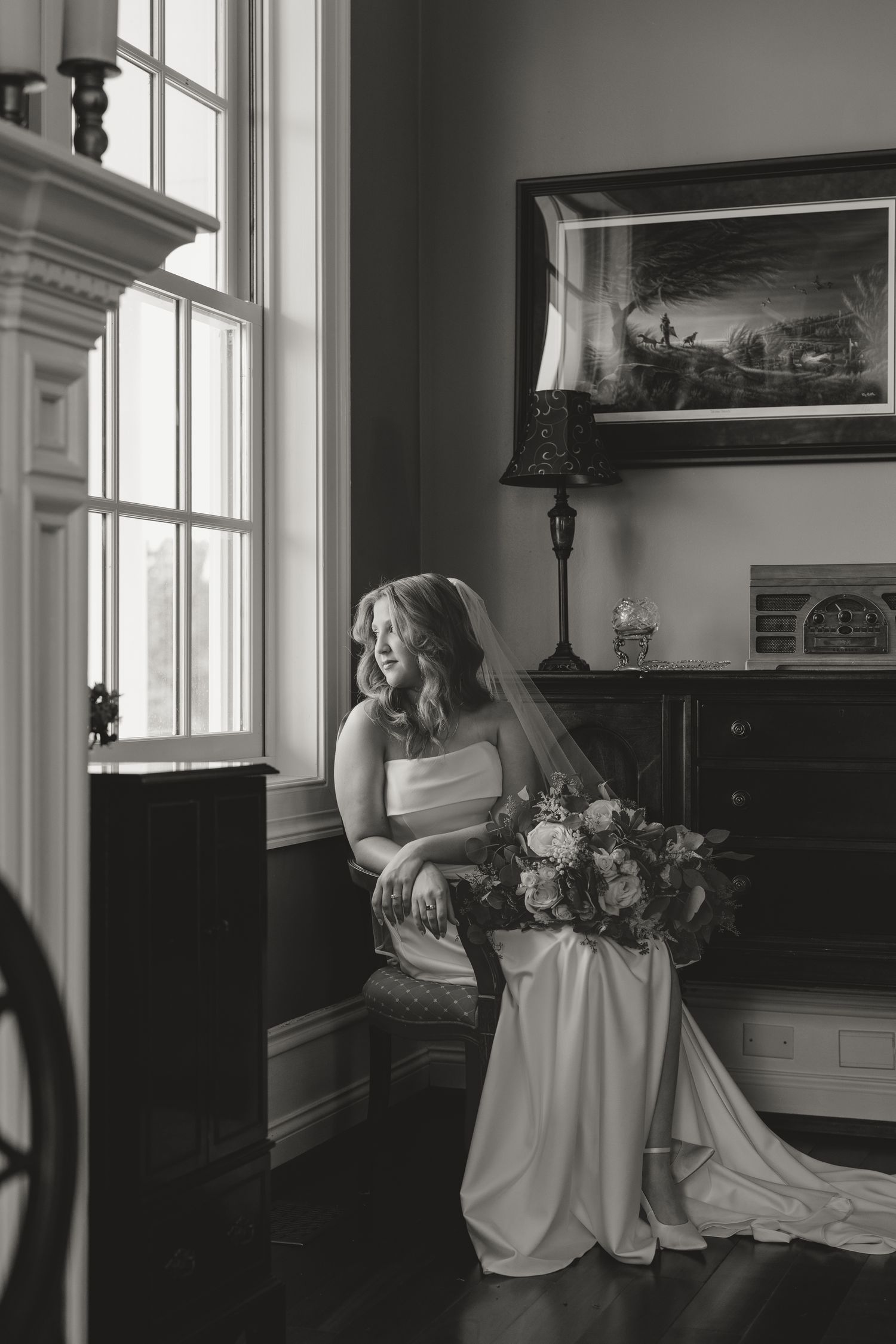 Black and white bridal portrait of a bride in a wedding gown holding a bouquet, seated by a window in an elegant room.