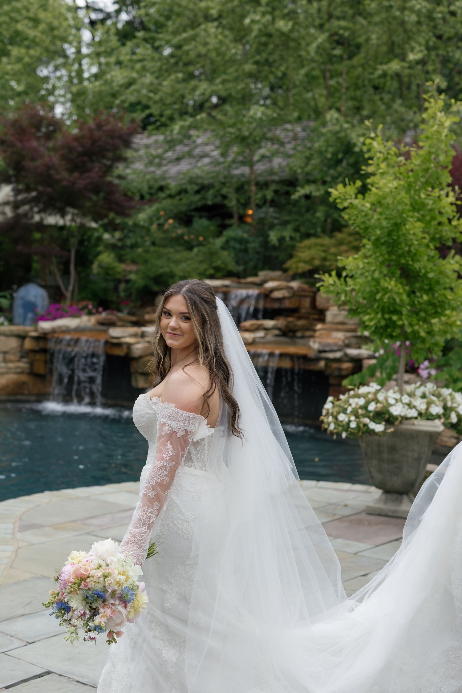 Bride in off-shoulder lace gown with flowing veil holds bouquet by stone waterfall garden.