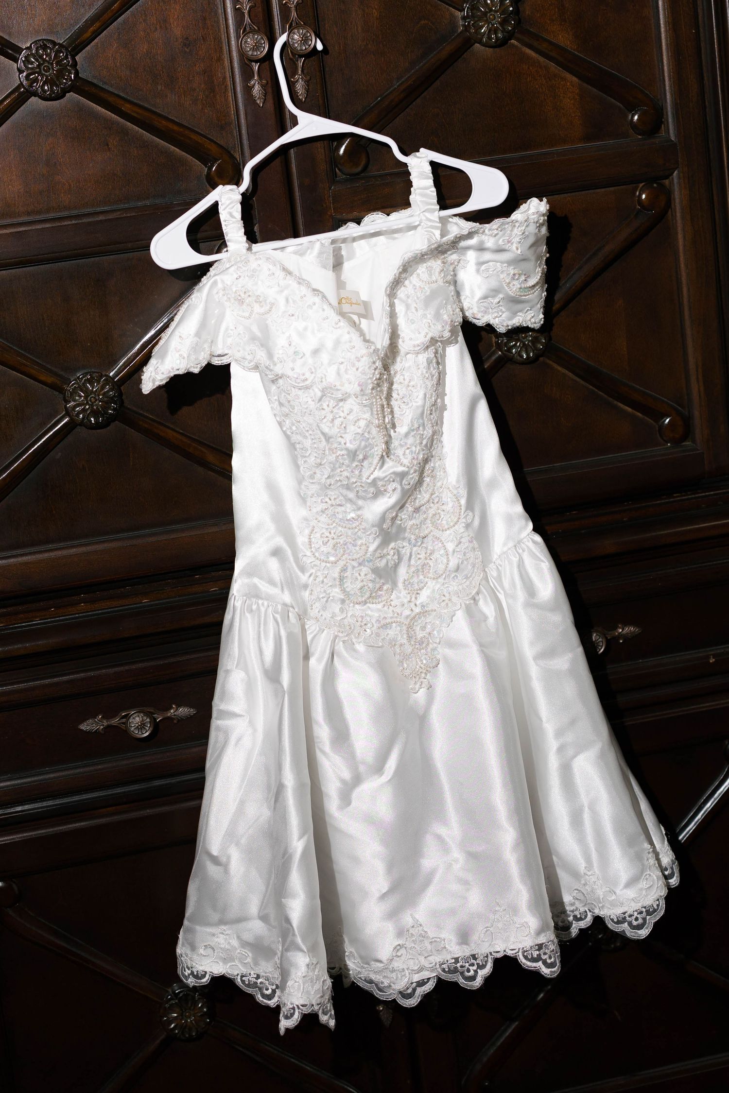 White lace off-shoulder flower girl dress hanging on hanger against dark wood cabinet.