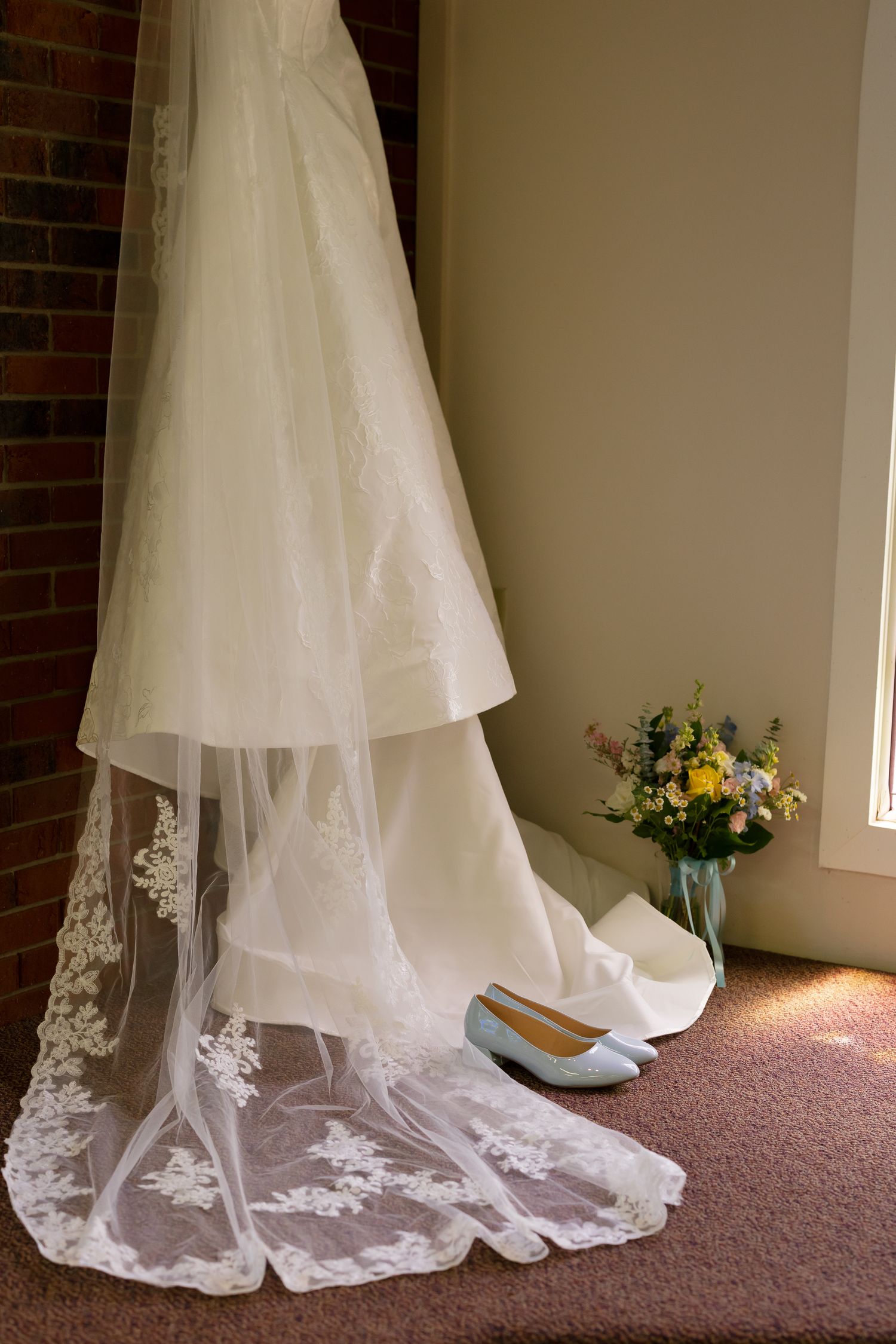 White lace wedding dress hanging near a window with blue bridal shoes and a colorful flower bouquet on the floor.