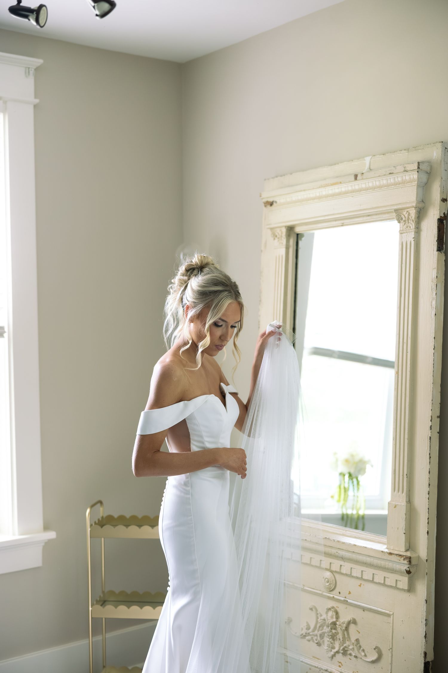 Bride in off-shoulder white gown holding veil beside ornate mirror in soft natural light.
