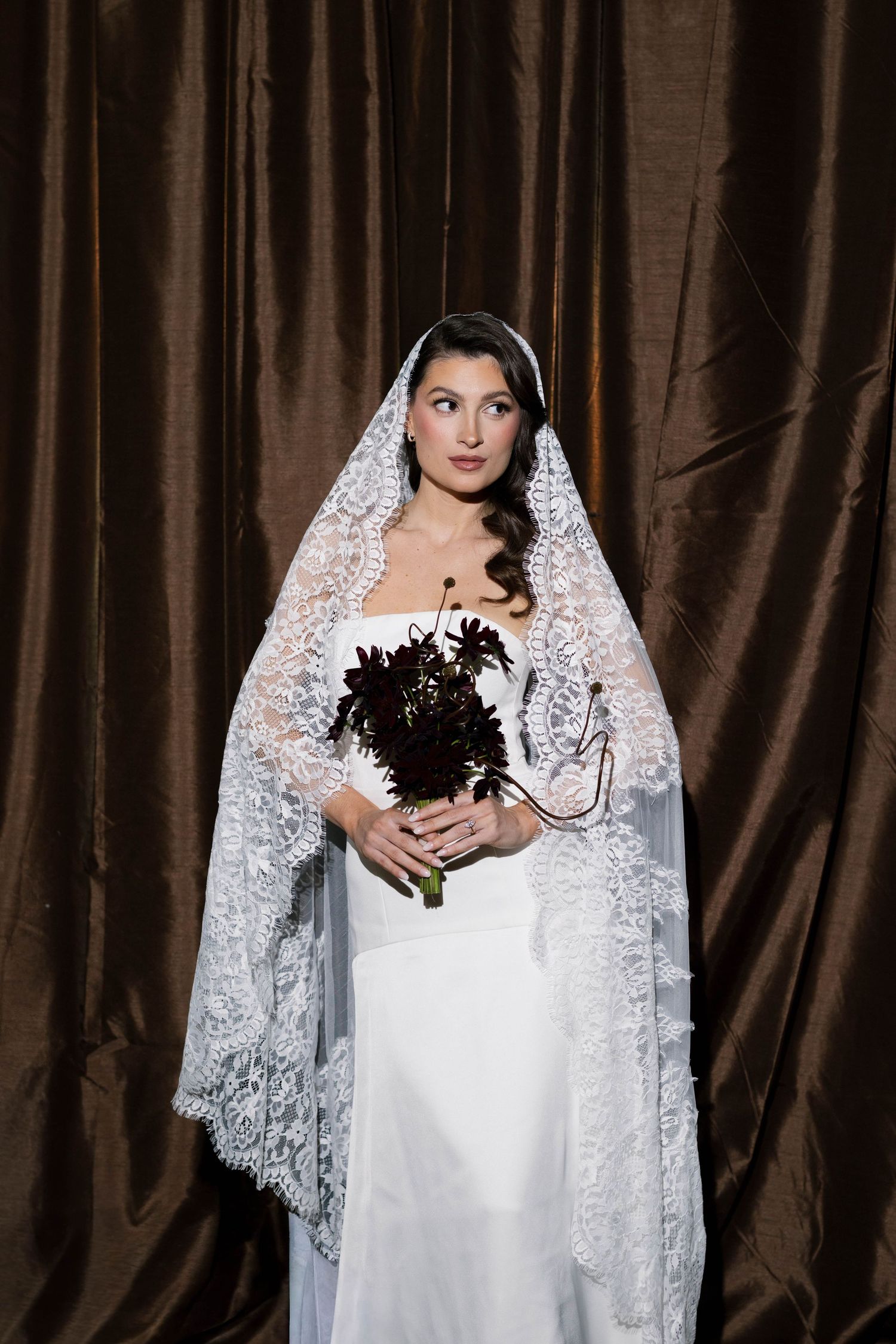 A dramatic bridal portrait featuring a white lace veil and dark bouquet against hanging brown drapery