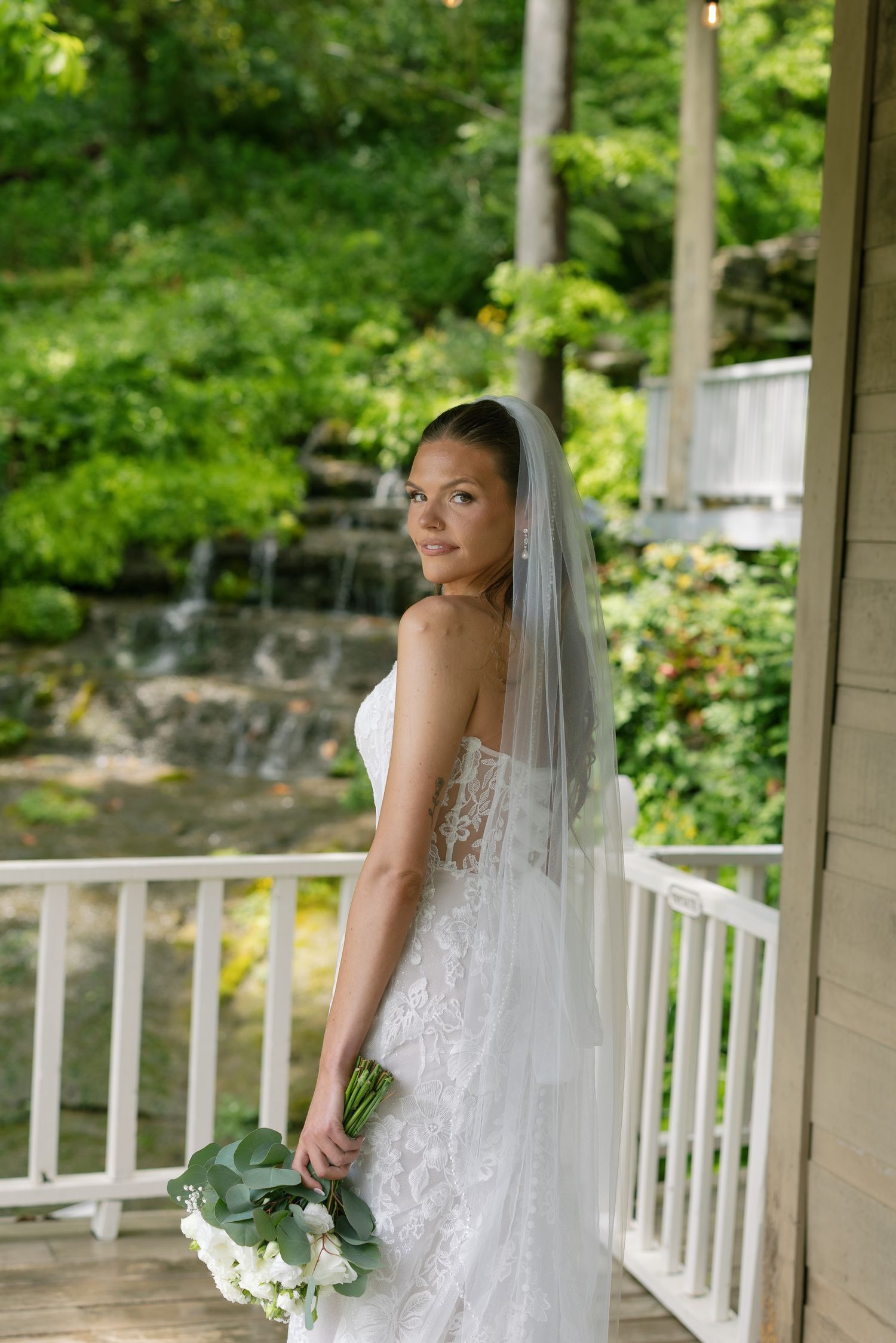 Bride in lace strapless gown and veil holds white bouquet on porch with waterfall backdrop.
