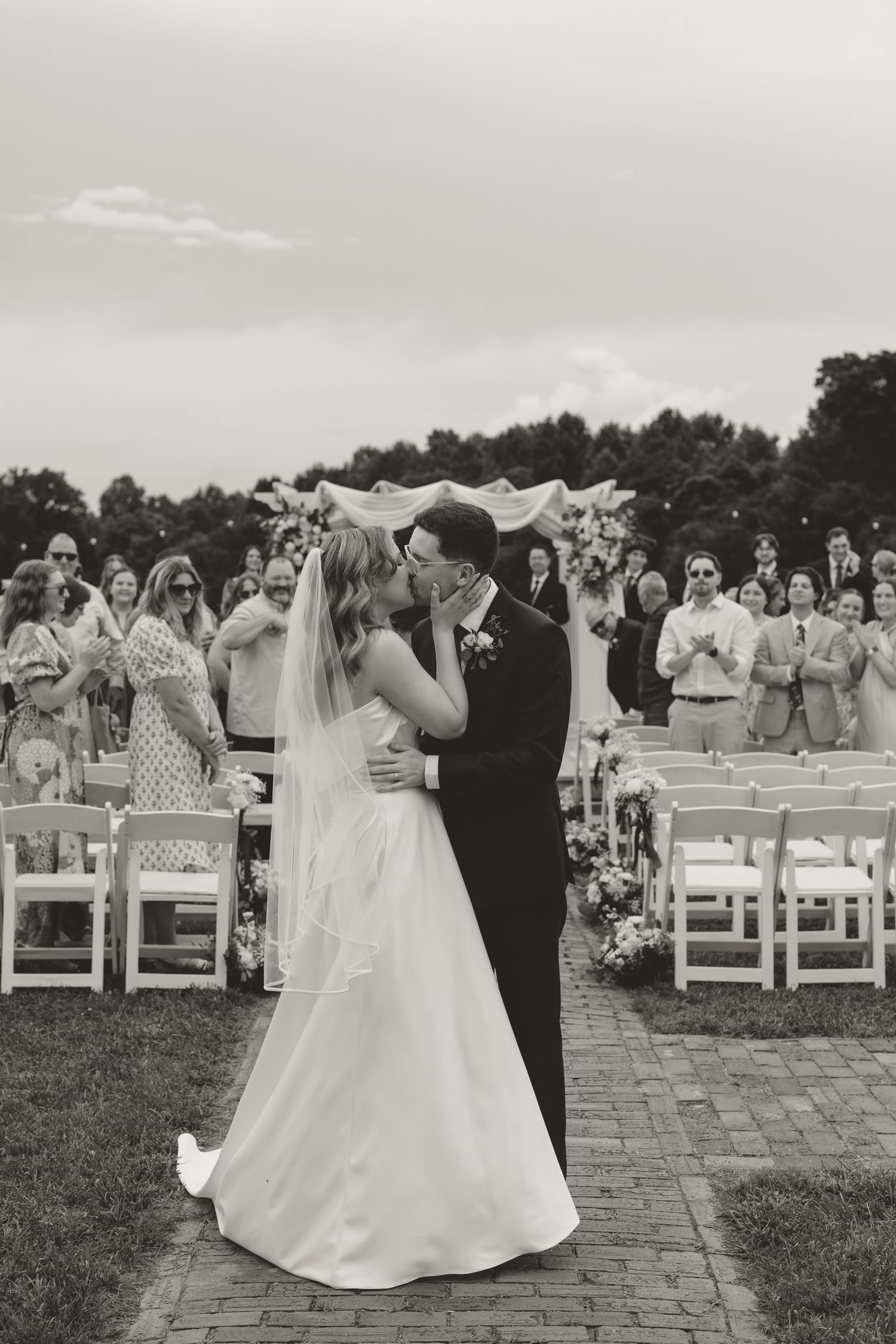 Black and white photo of a bride and groom sharing their first kiss at an outdoor wedding ceremony with guests applauding.