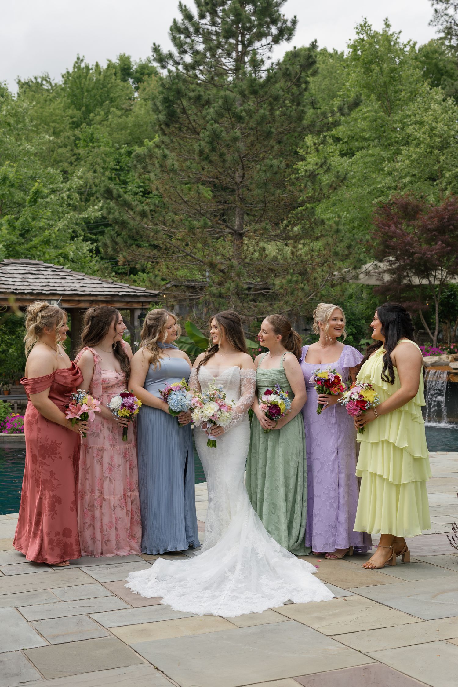 Bride in white gown poses with six bridesmaids in colorful mismatched dresses holding bouquets outdoors near a pool.