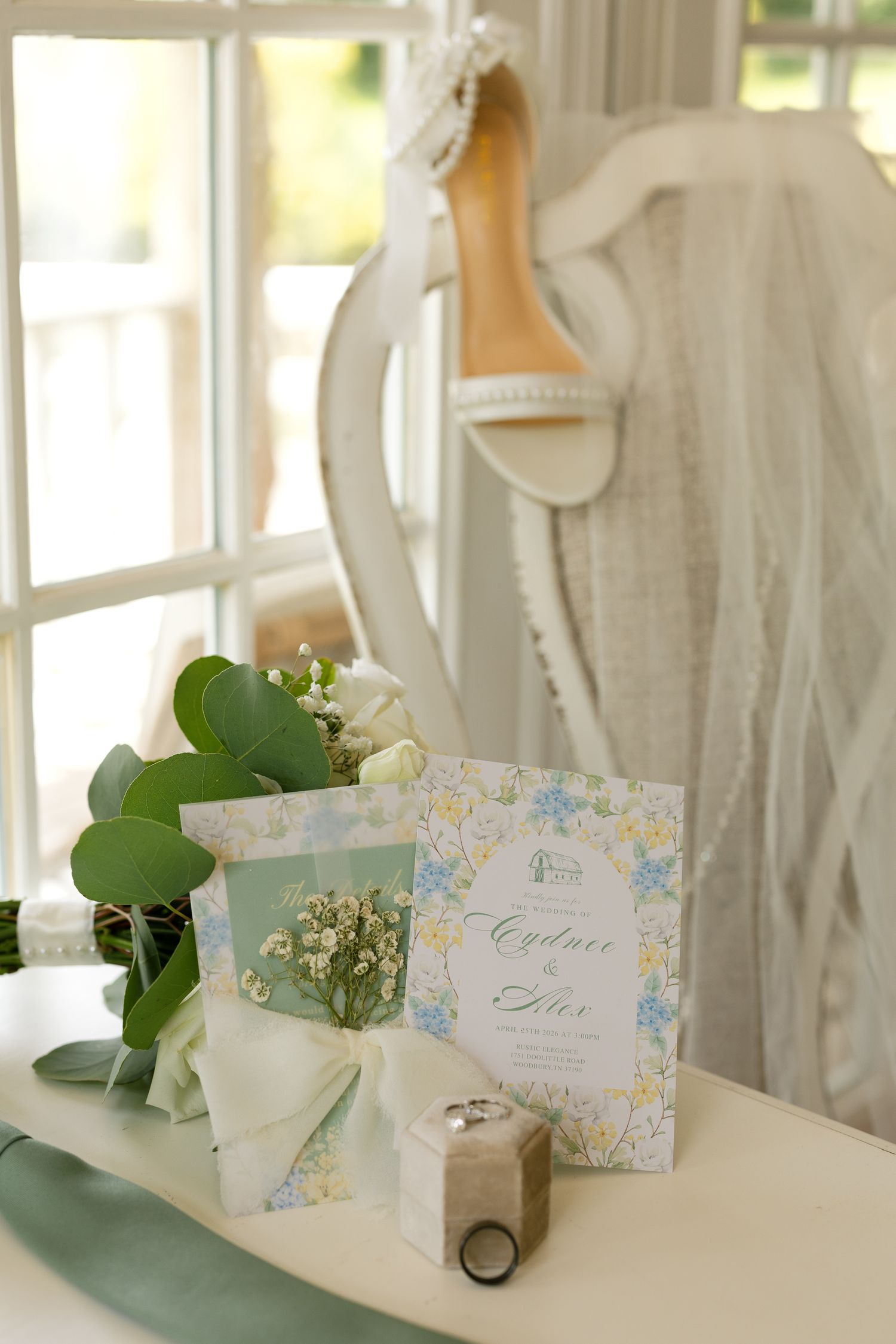 Elegant wedding flat lay with floral invitation, bridal bouquet, ring box, and white heels on a vintage chair.
