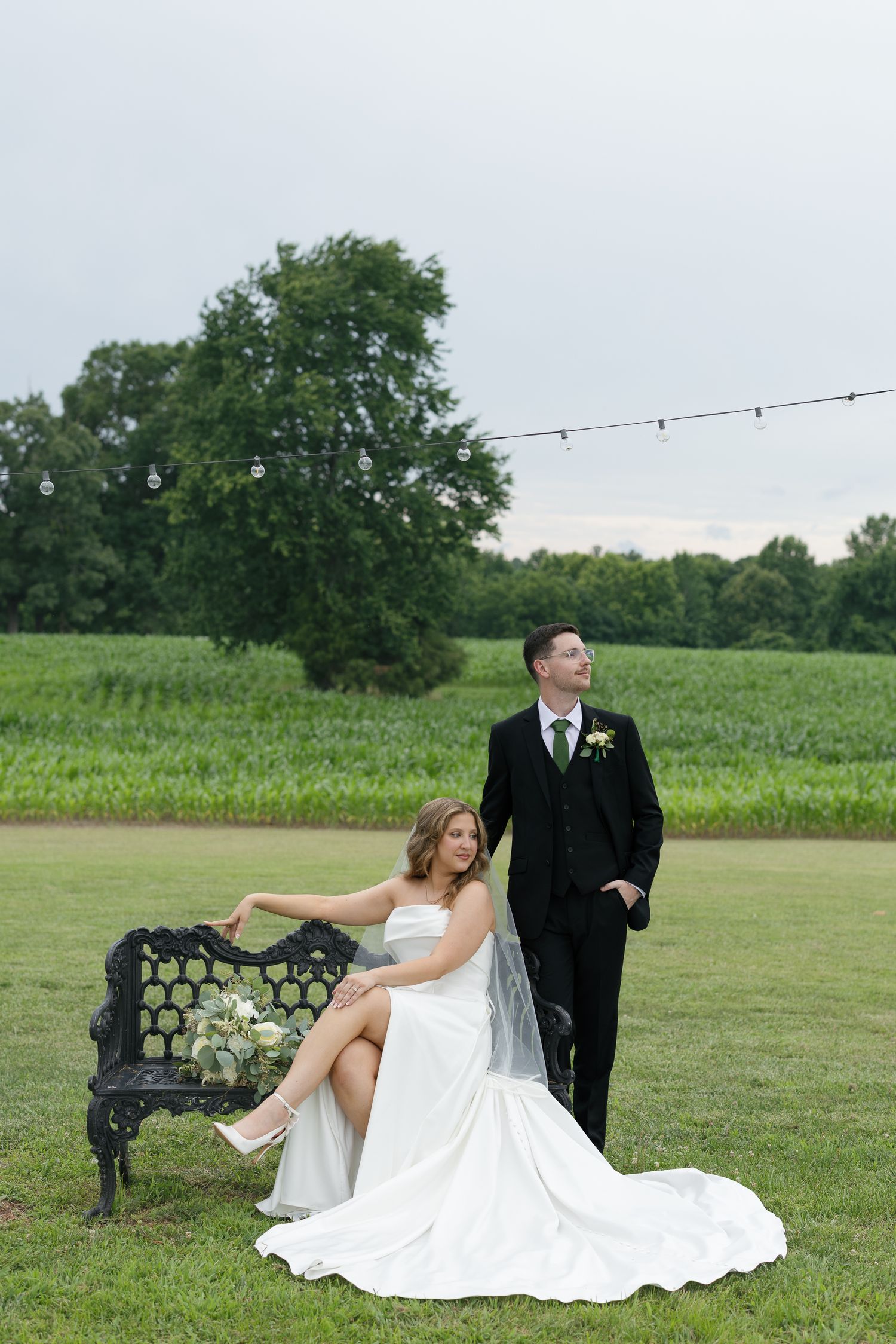 Bride in white gown sits on ornate bench beside groom in black suit, posing in lush green field with string lights.