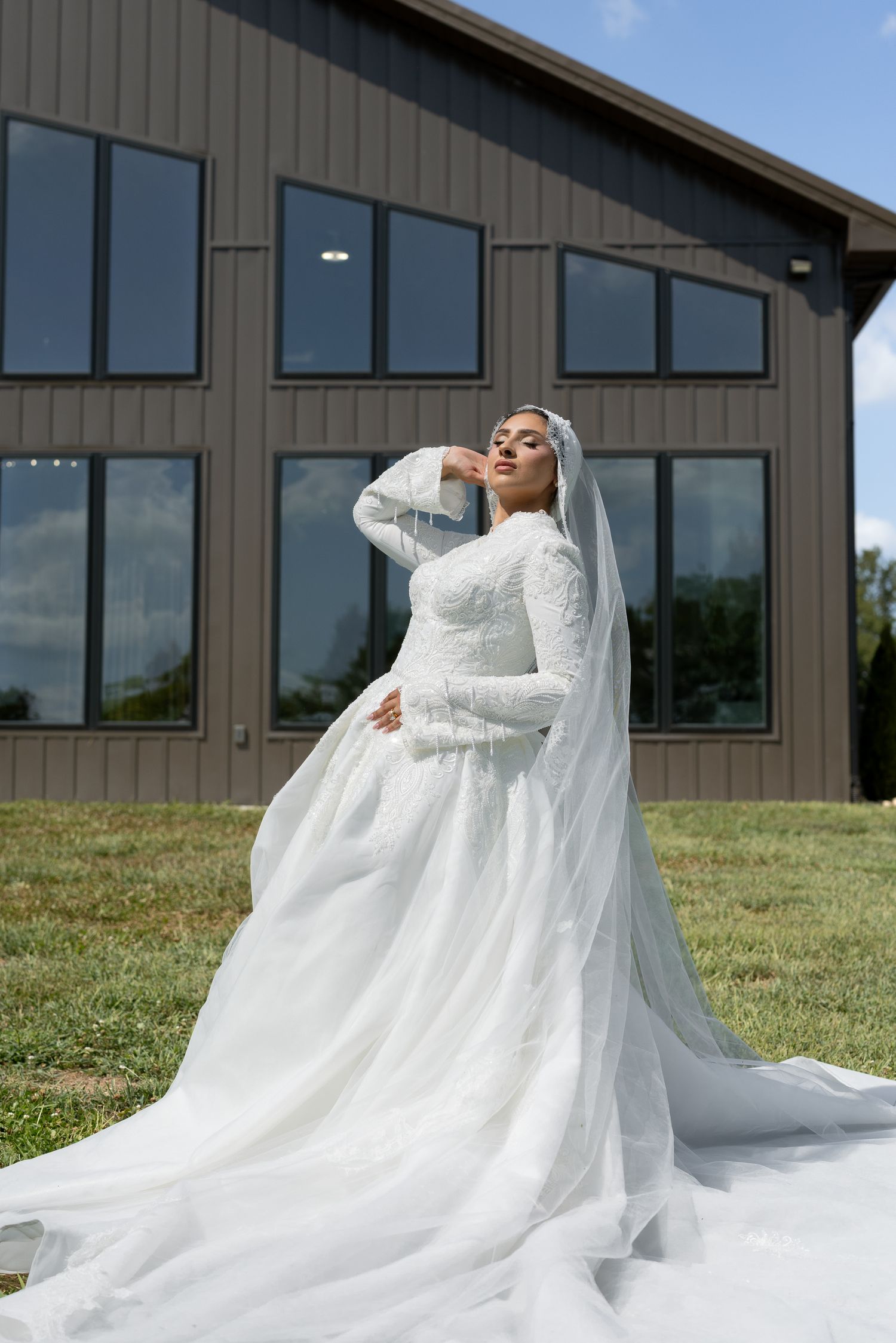 Bride in elegant long-sleeve white ballgown with veil poses outdoors before a modern dark barn venue.