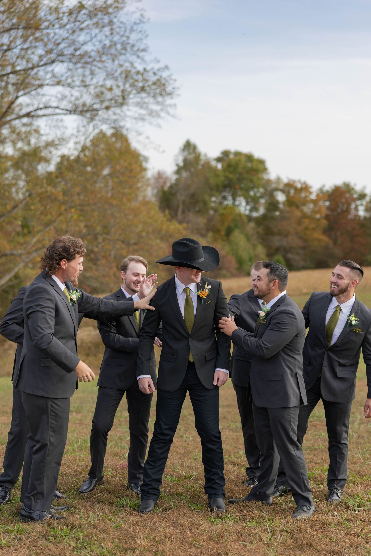 Kentucky country Groomsmen in dark suits with gold ties laugh outdoors in a fall field, one wearing a black cowboy hat.