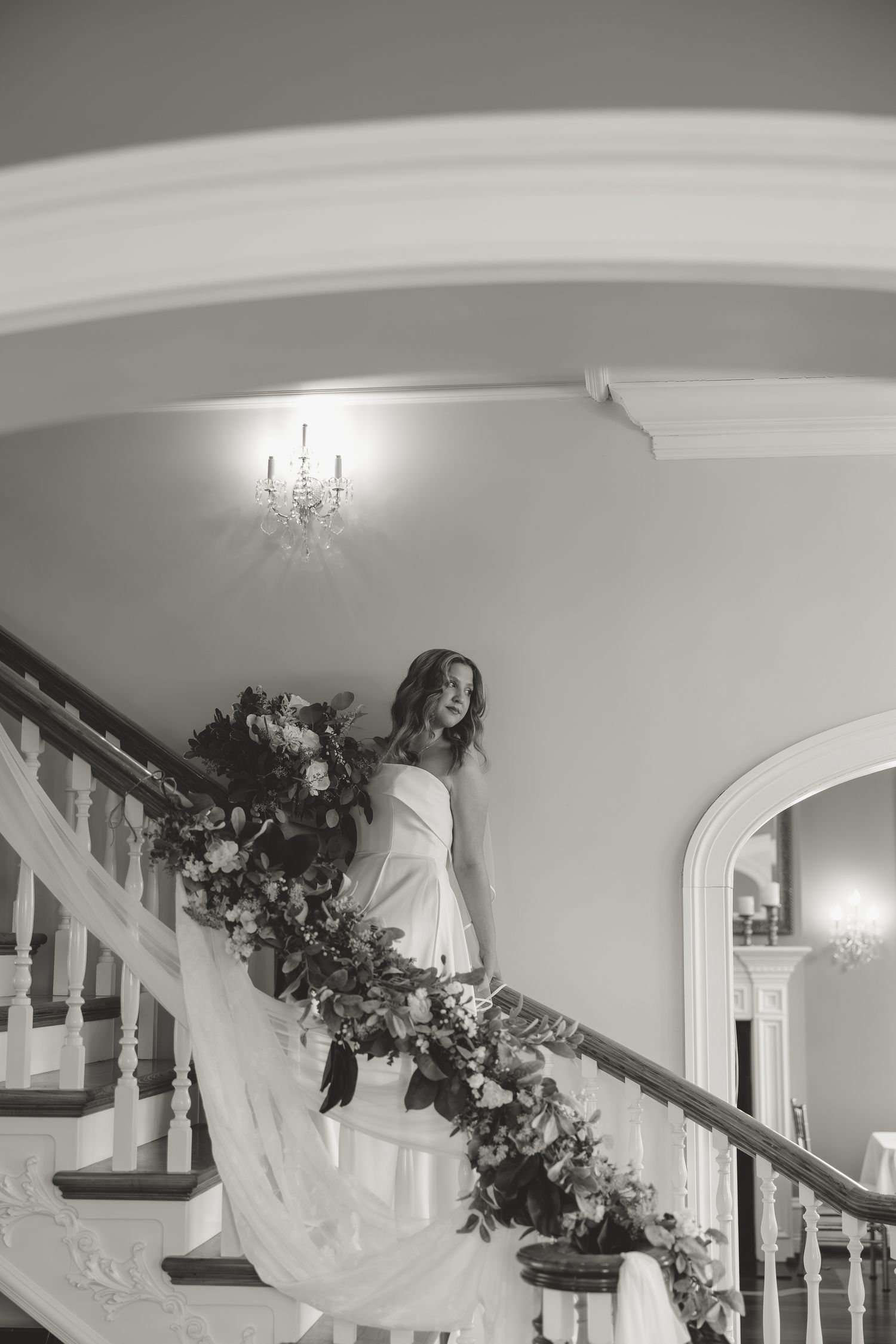 Bride in white gown descends floral-adorned staircase in black and white wedding photo.