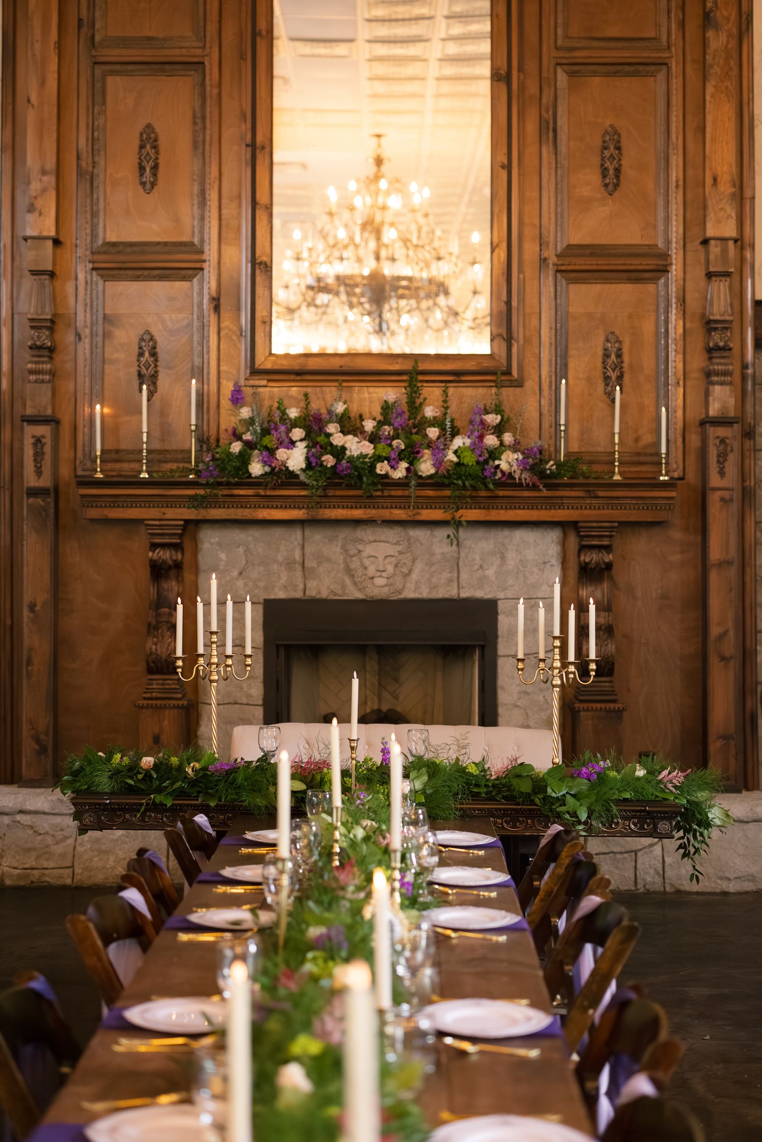 Elegant wedding reception table with candles, greenery garland, and floral fireplace mantel in a wood-paneled hall.