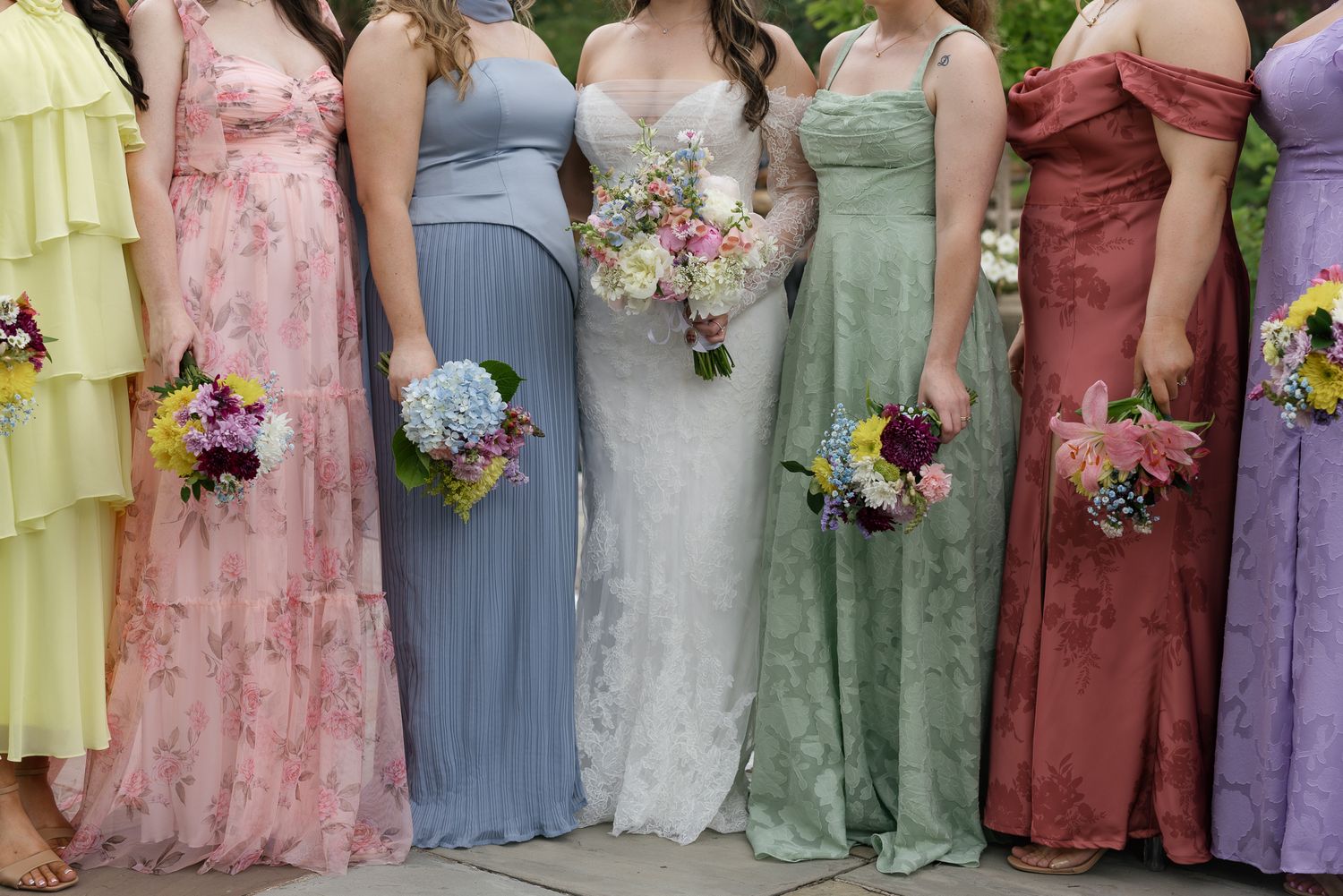 Bride in white lace gown stands with colorful bridesmaids in pastel dresses holding floral bouquets outdoors.