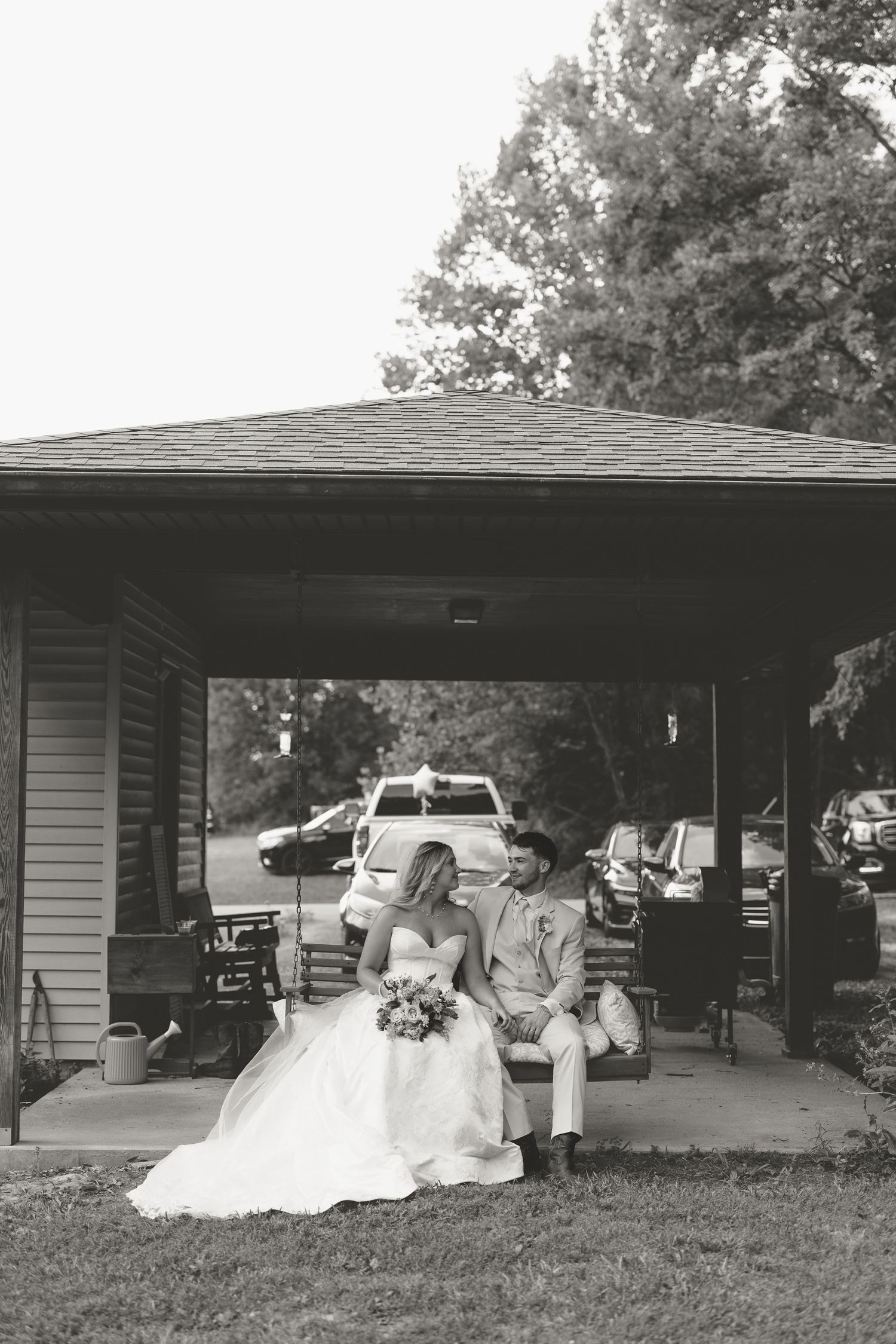 Black and white photo of a bride and groom sitting together on a porch swing, bride holding a bouquet.