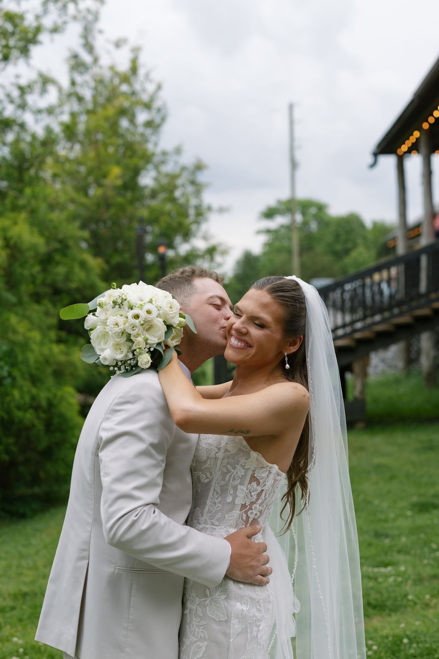 Groom in gray suit kisses bride in white lace gown and veil, holding white bouquet, outdoors at wedding venue.