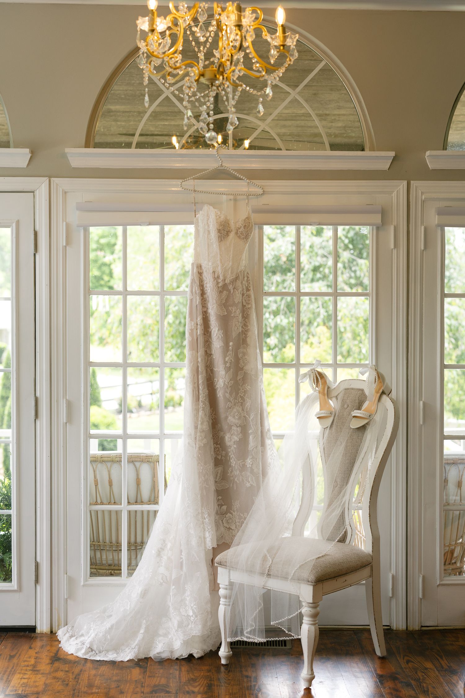 Elegant lace wedding dress hanging in a sunlit room with a crystal chandelier and white vintage chair.