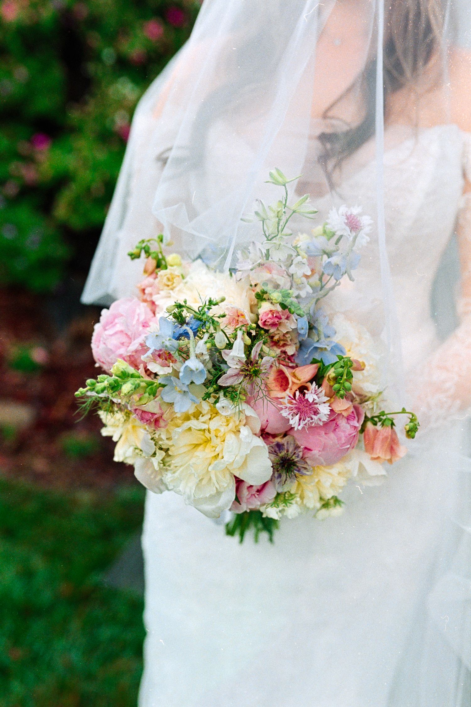 Bride in white lace gown holding a pastel wedding bouquet with pink peonies, blue delphinium, and mixed spring flowers.