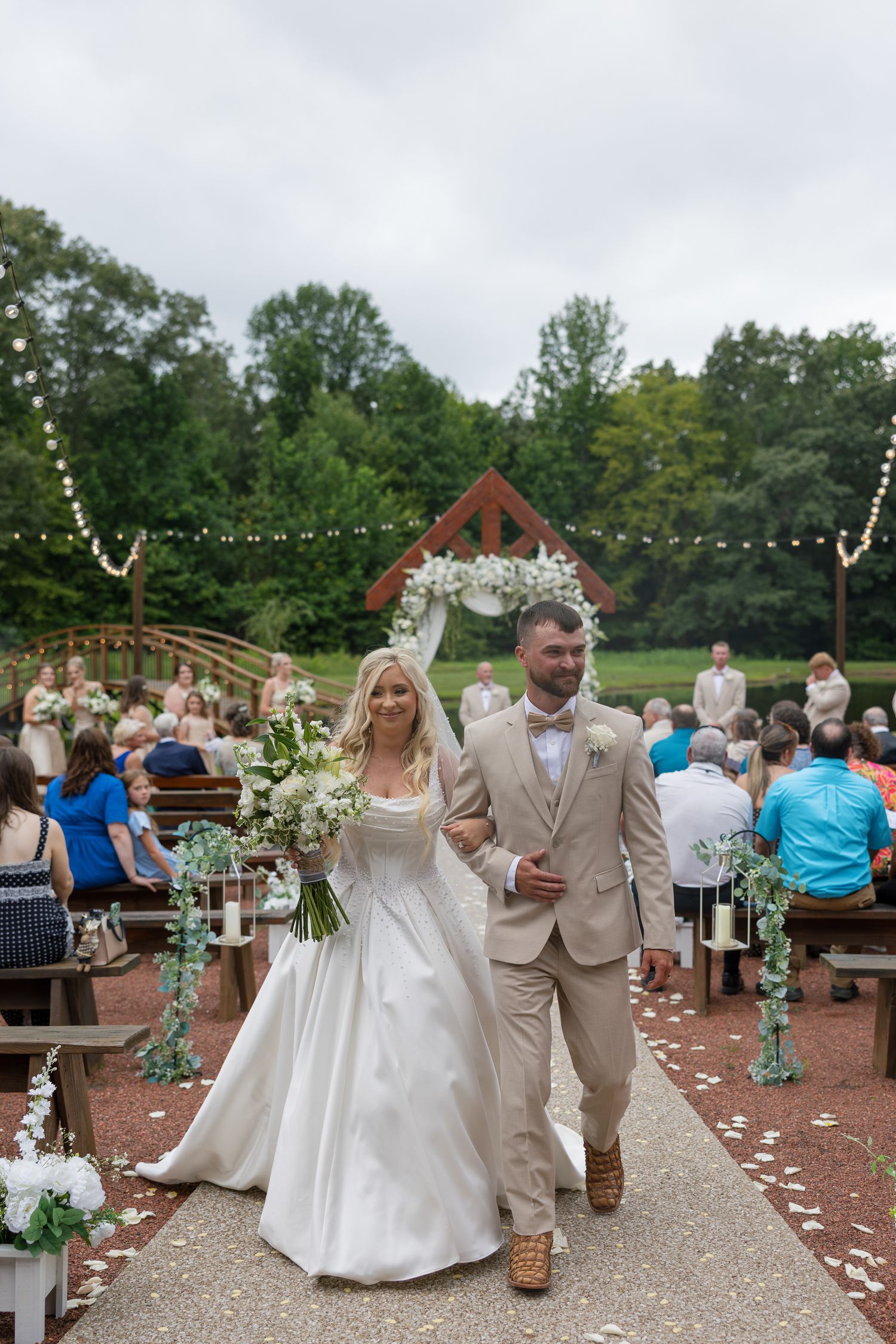 Bride and groom walk down outdoor aisle after wedding ceremony, surrounded by guests and floral arch backdrop.