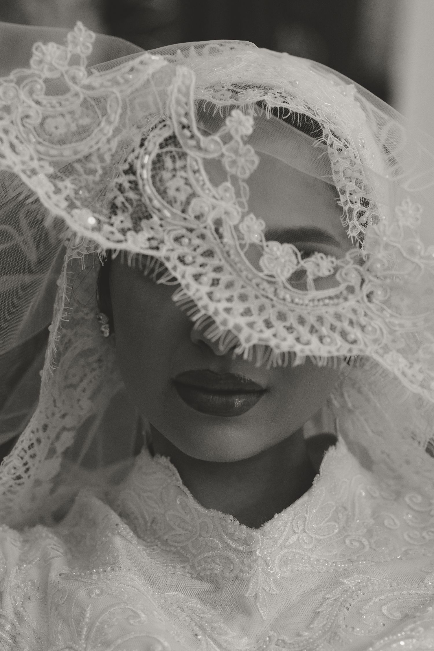 Black and white portrait of a bride wearing intricate lace veil covering her eyes, elegant bridal fashion photography.