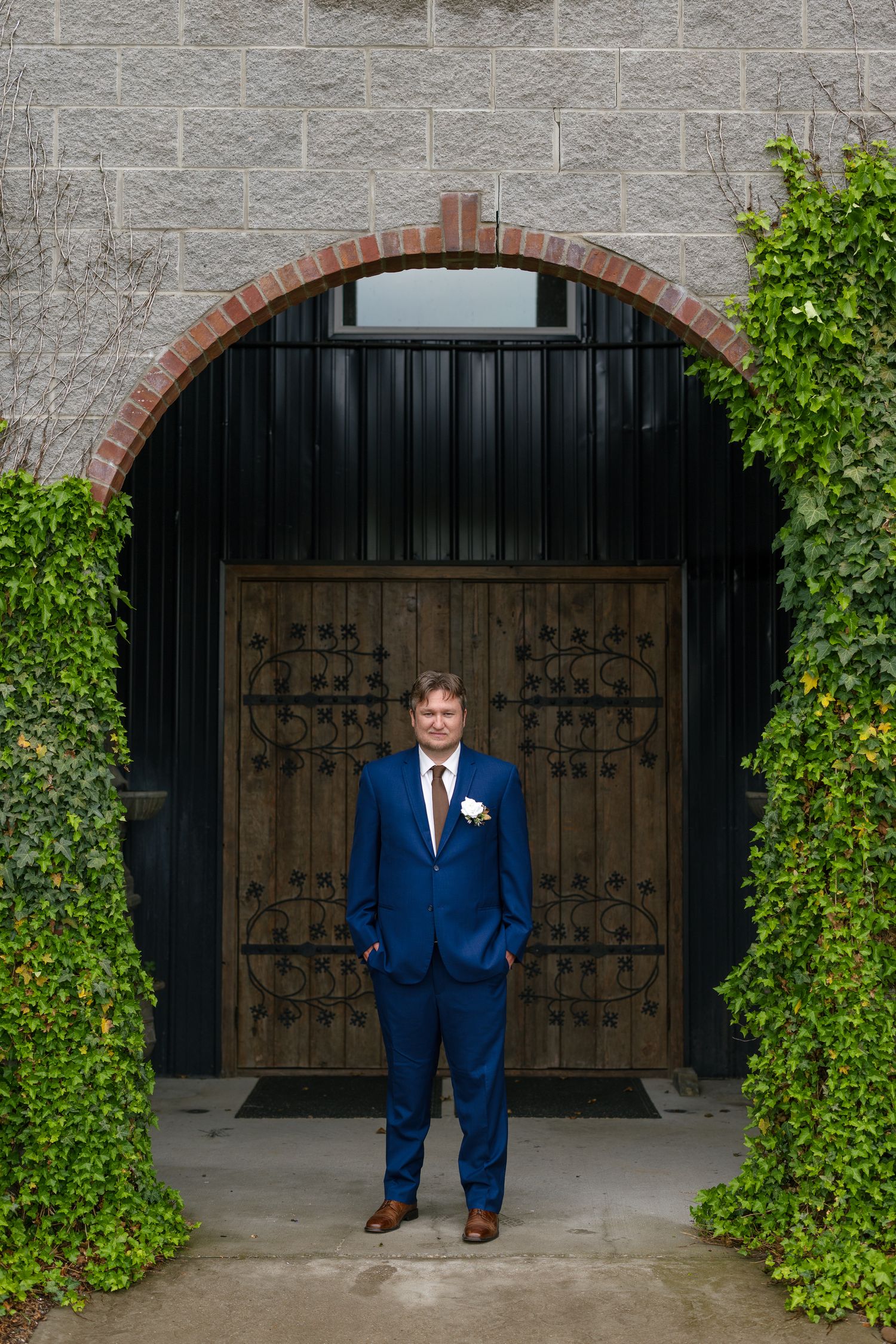 Groom in navy blue suit with boutonniere stands before ornate wooden doors framed by ivy and brick arch.