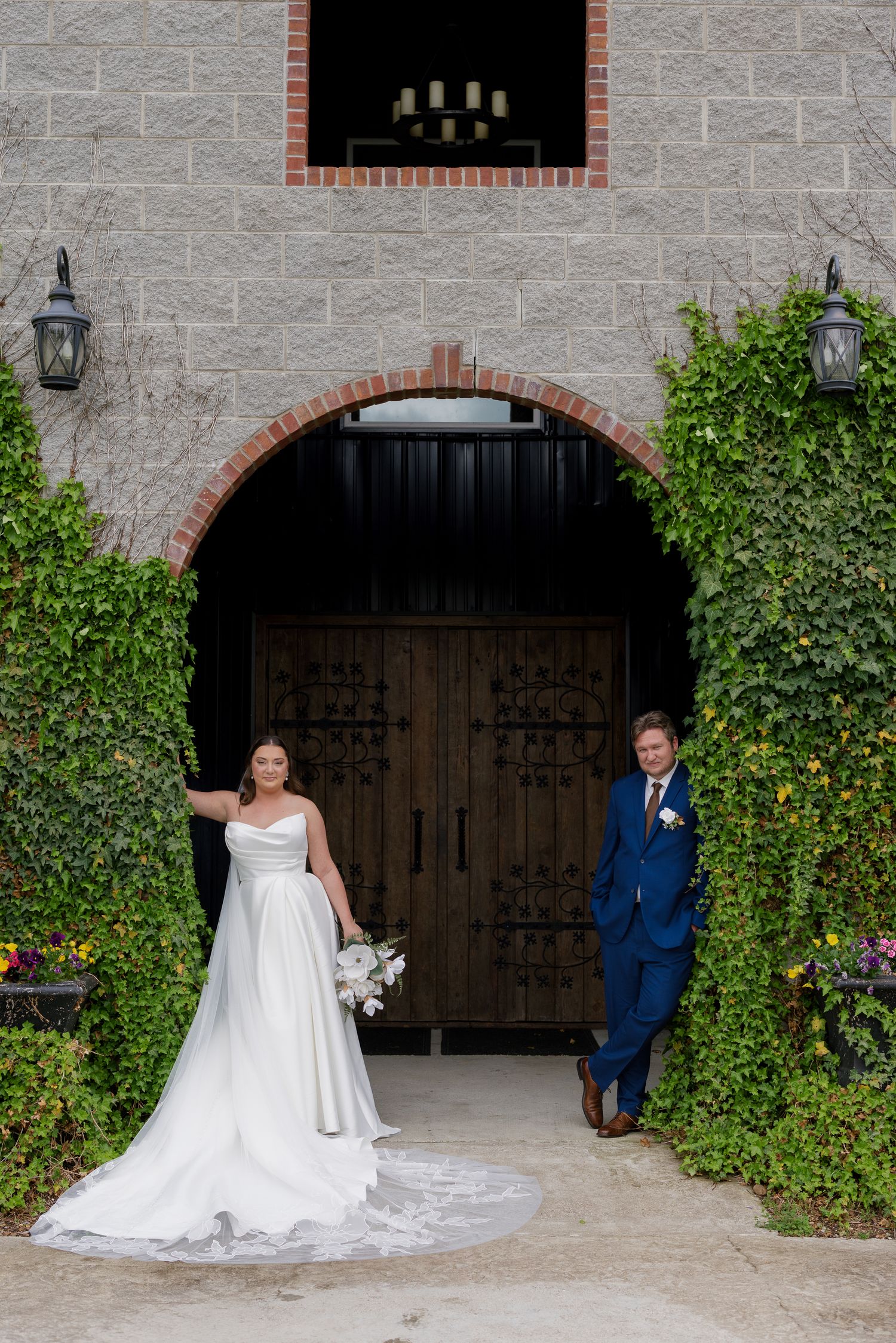 Bride in white gown and groom in blue suit pose under ivy-covered stone arch with ornate wooden doors.
