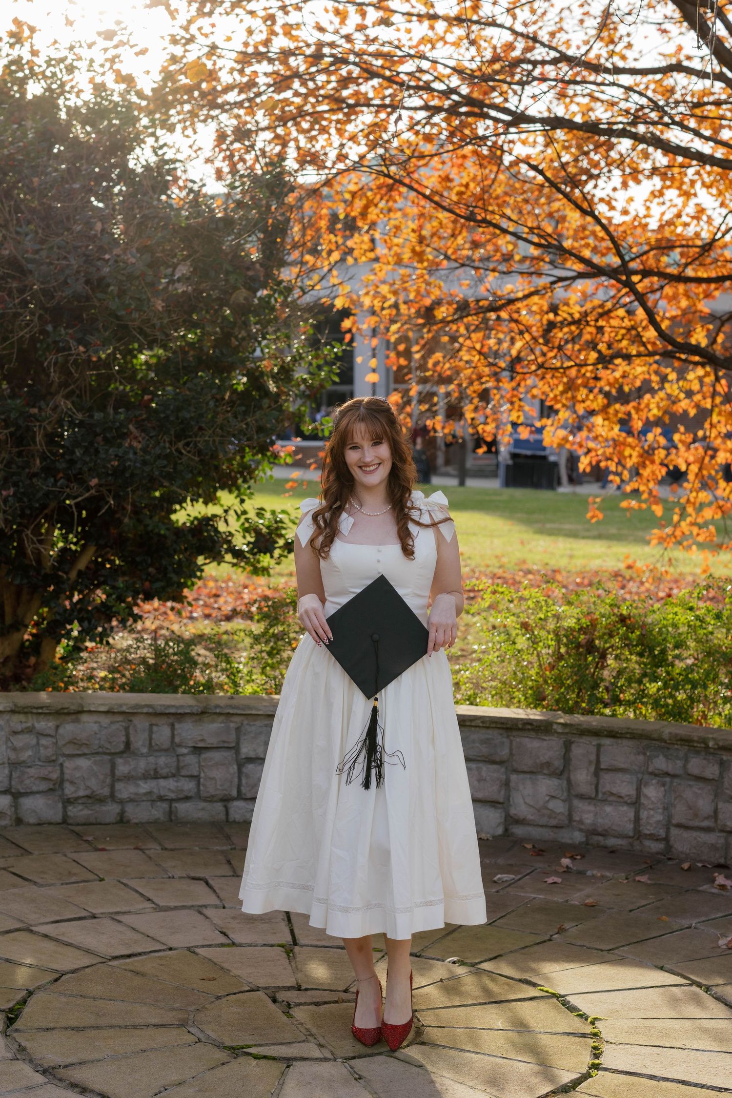 A college graduate in a white dress and red shoes standing on a stone patio with autumn foliage overhead on a sunny fall day at Belmont