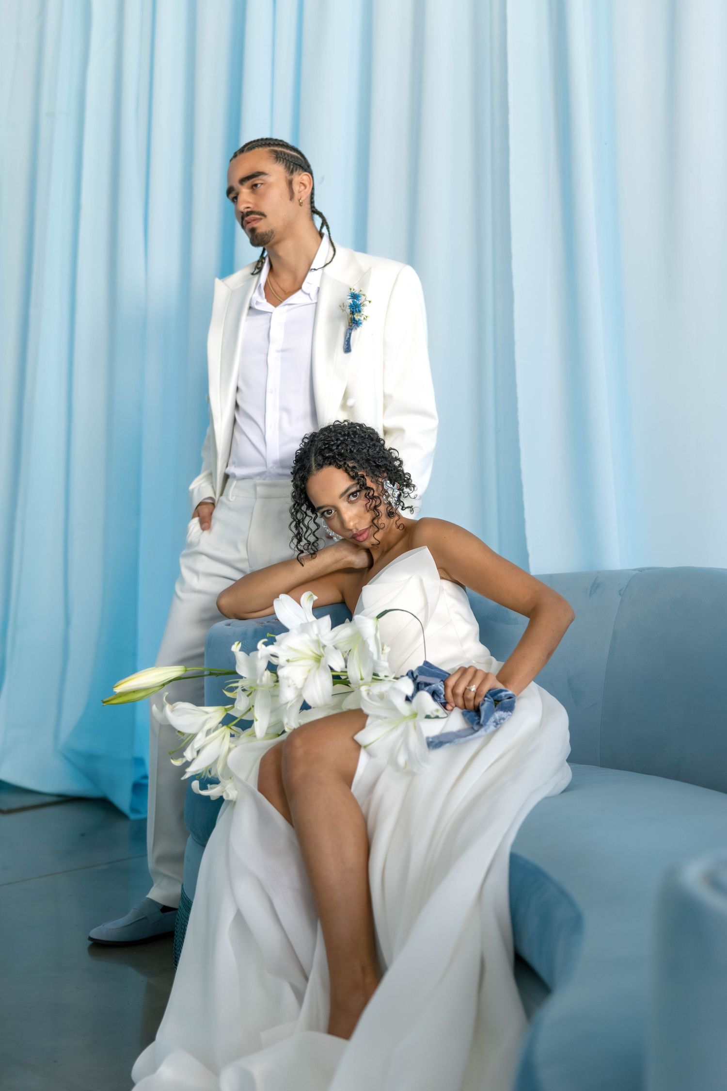 Romantic wedding couple in white outfits sitting on blue couch with lilies and baby blue drapery