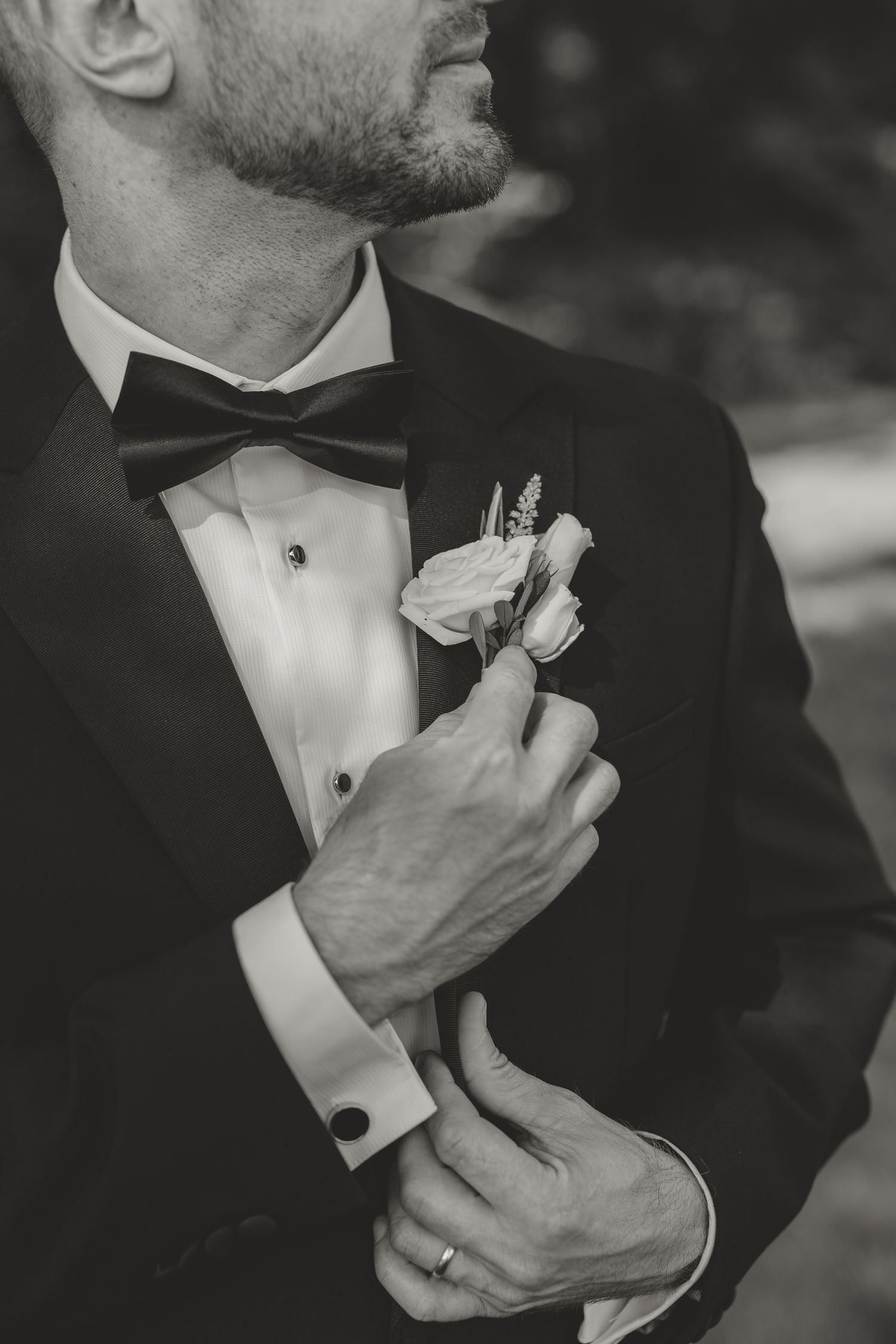 Black and white photo of a groom in a tuxedo with bow tie adjusting his floral boutonniere, wearing a wedding ring.
