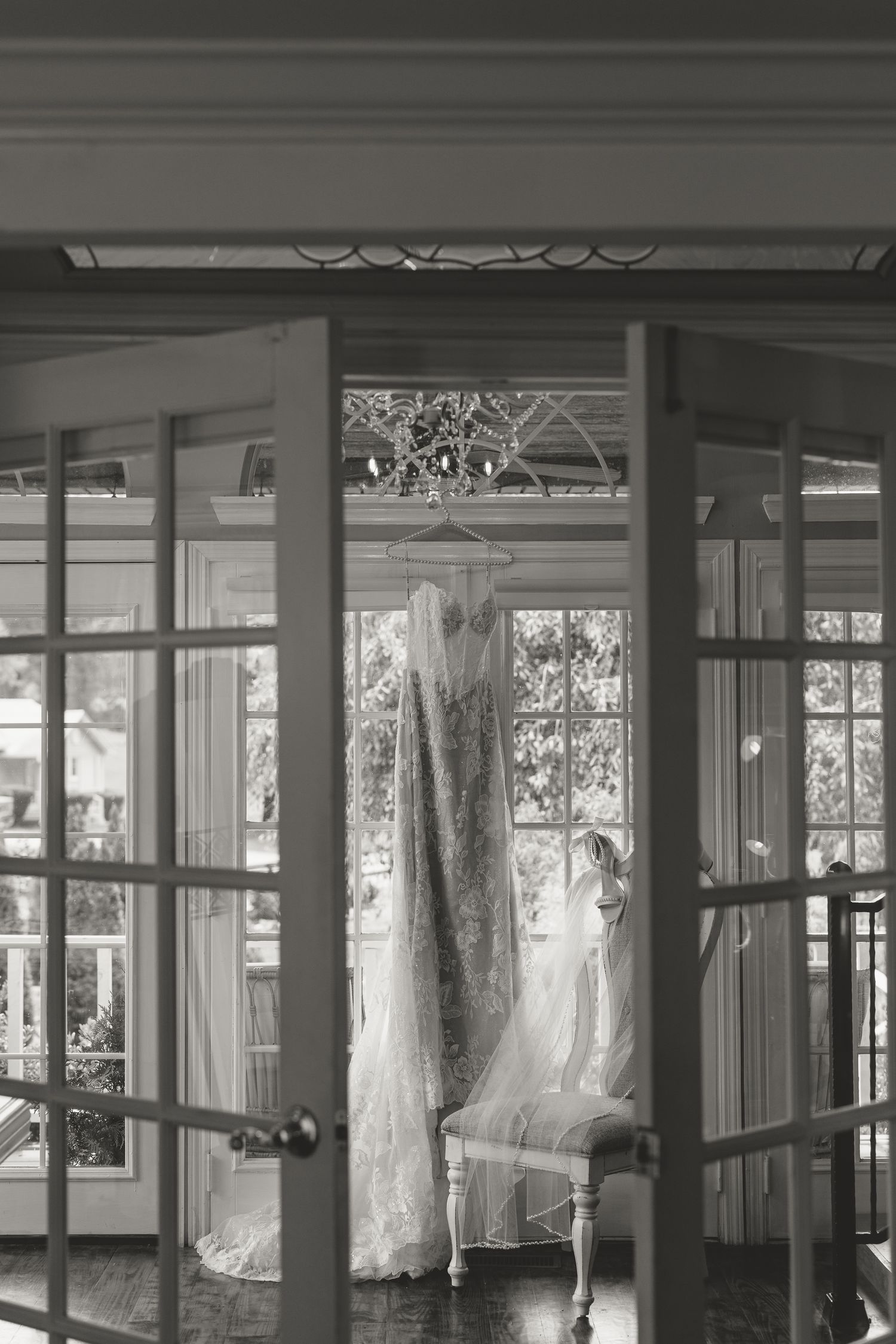 Black and white photo of an elegant wedding dress hanging in sunlit French doors of a classic interior.