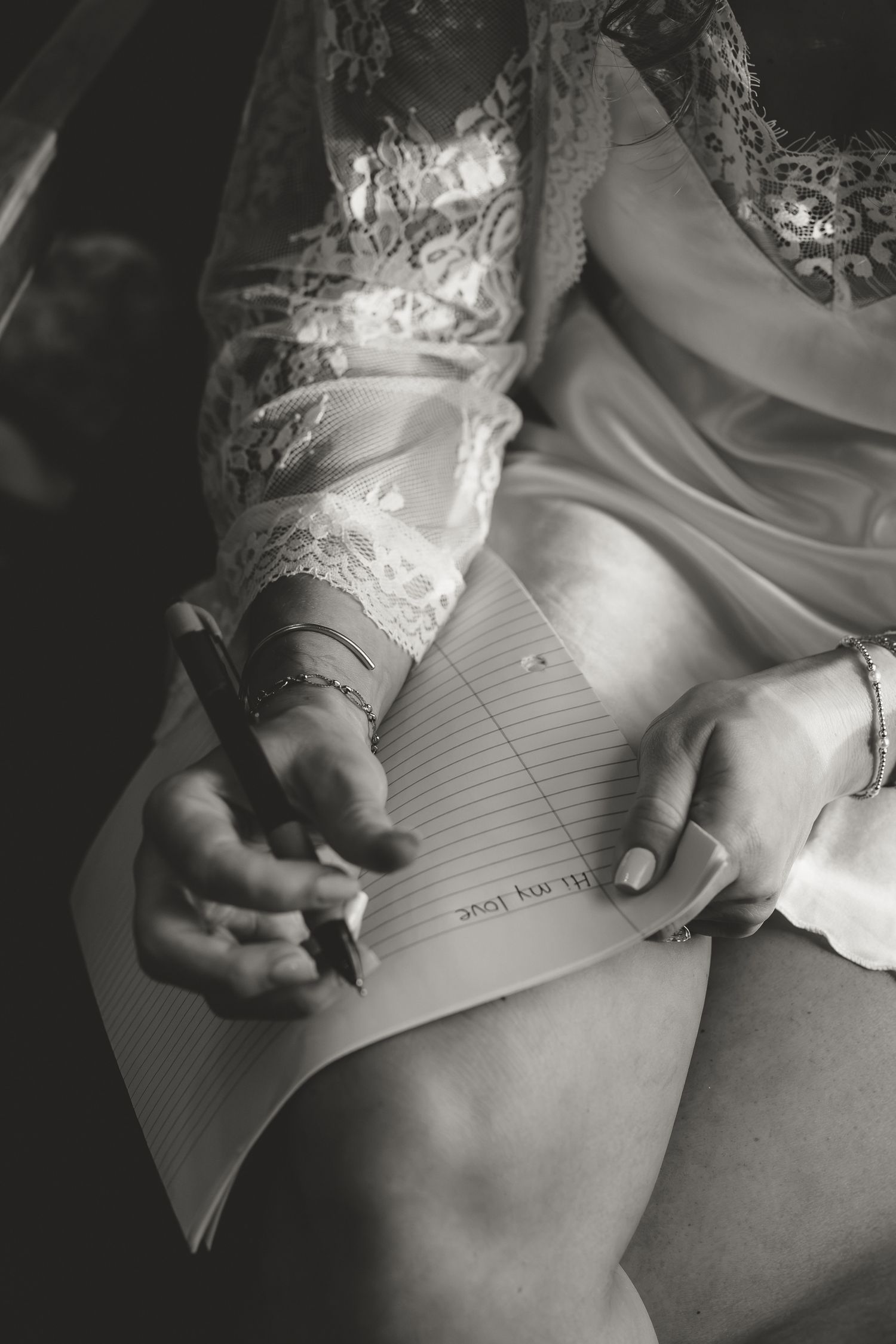 Black and white photo of a bride in lace robe writing a heartfelt letter with a pen on lined paper.