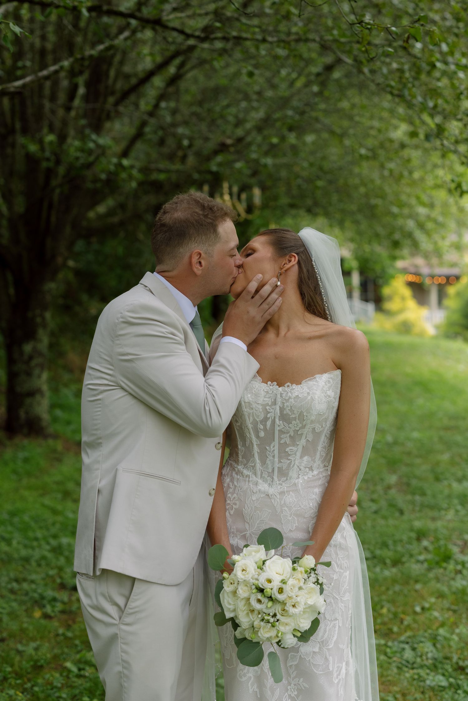Bride and groom share a romantic kiss outdoors, she holds white bouquet, wearing lace gown and veil, he in beige suit.