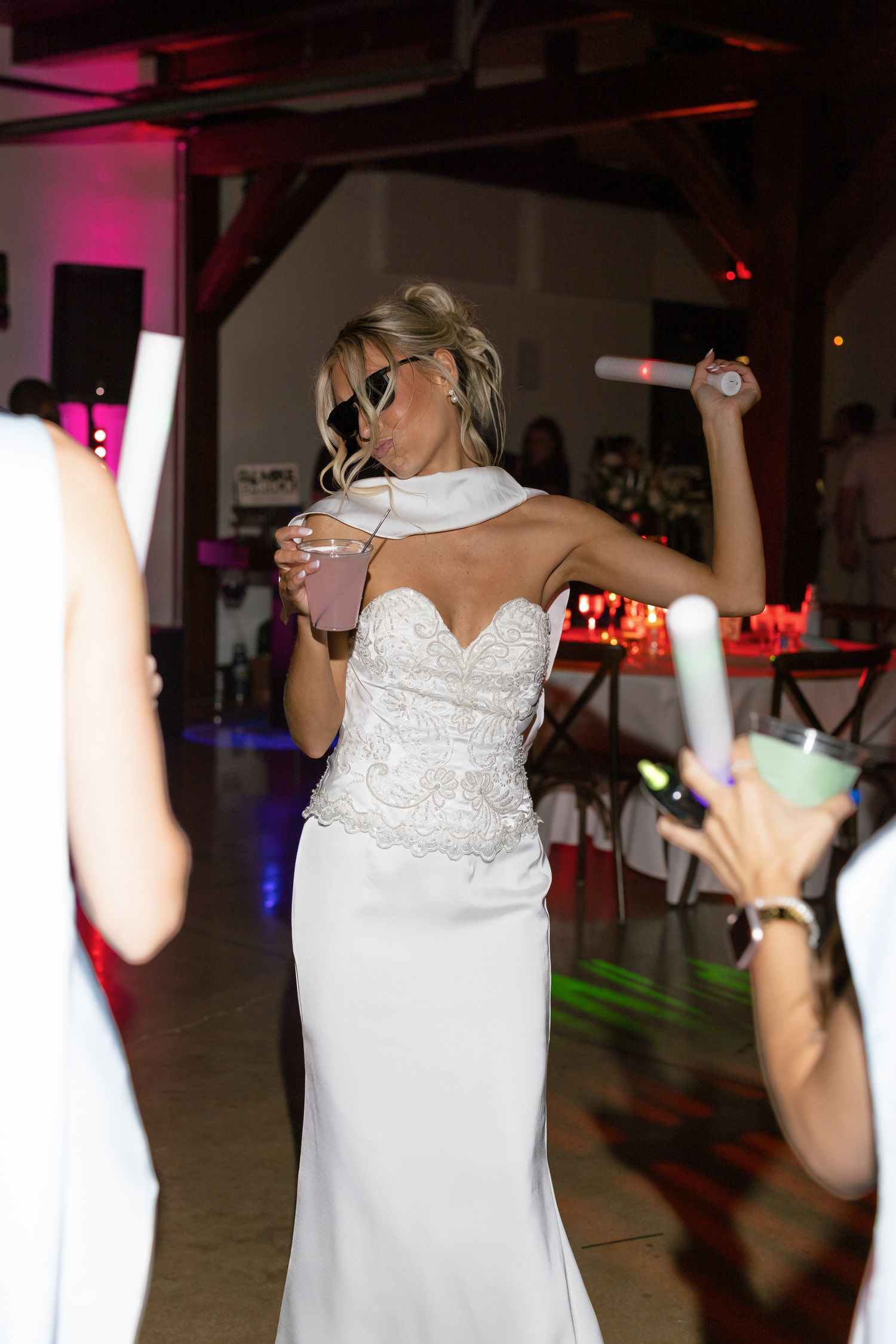 Bride in white lace gown dancing at wedding reception, sipping drink, wearing sunglasses, surrounded by guests with glow sticks.