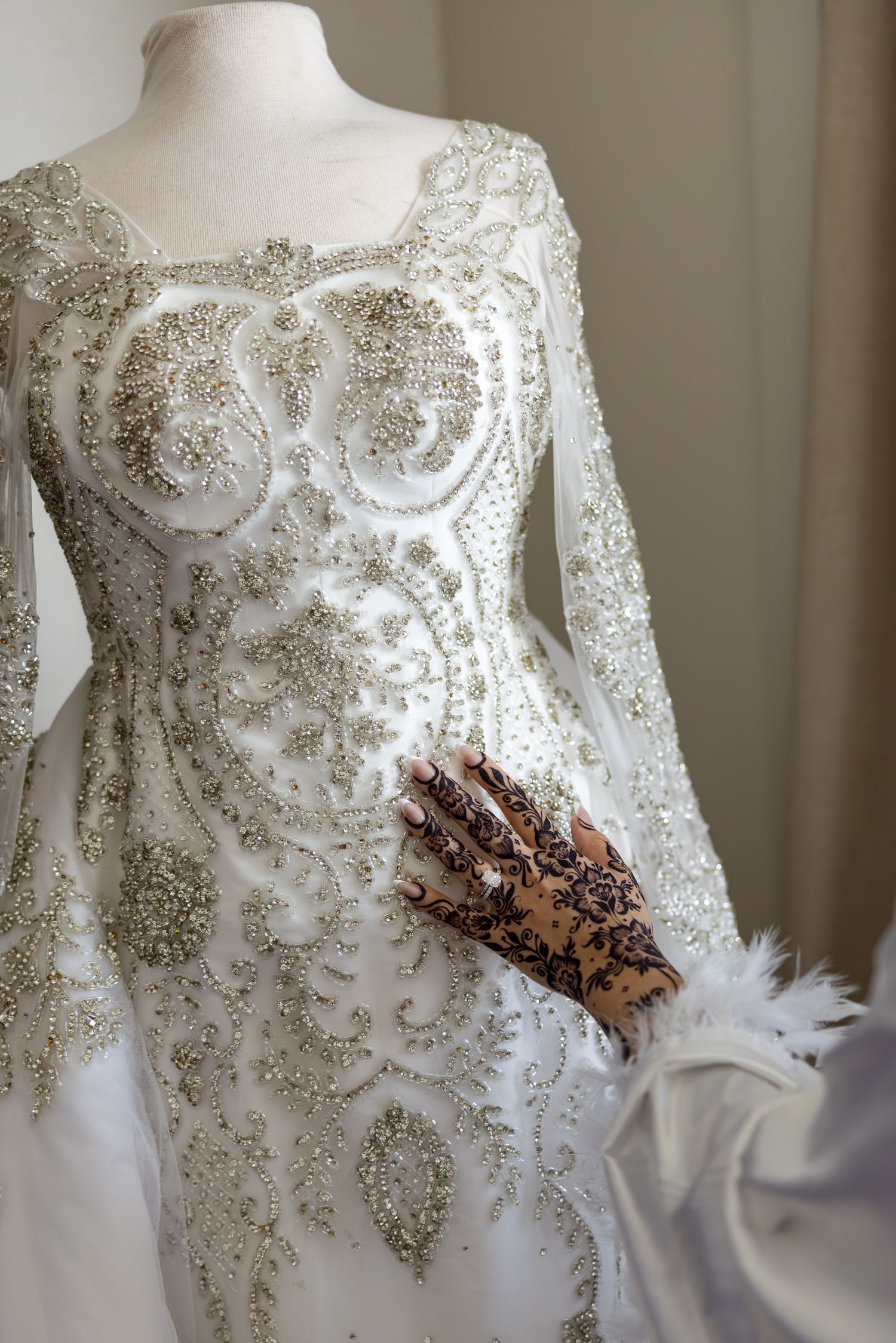 Ornate white bridal gown with silver beading and a henna-adorned hand, featuring feather cuff detail.