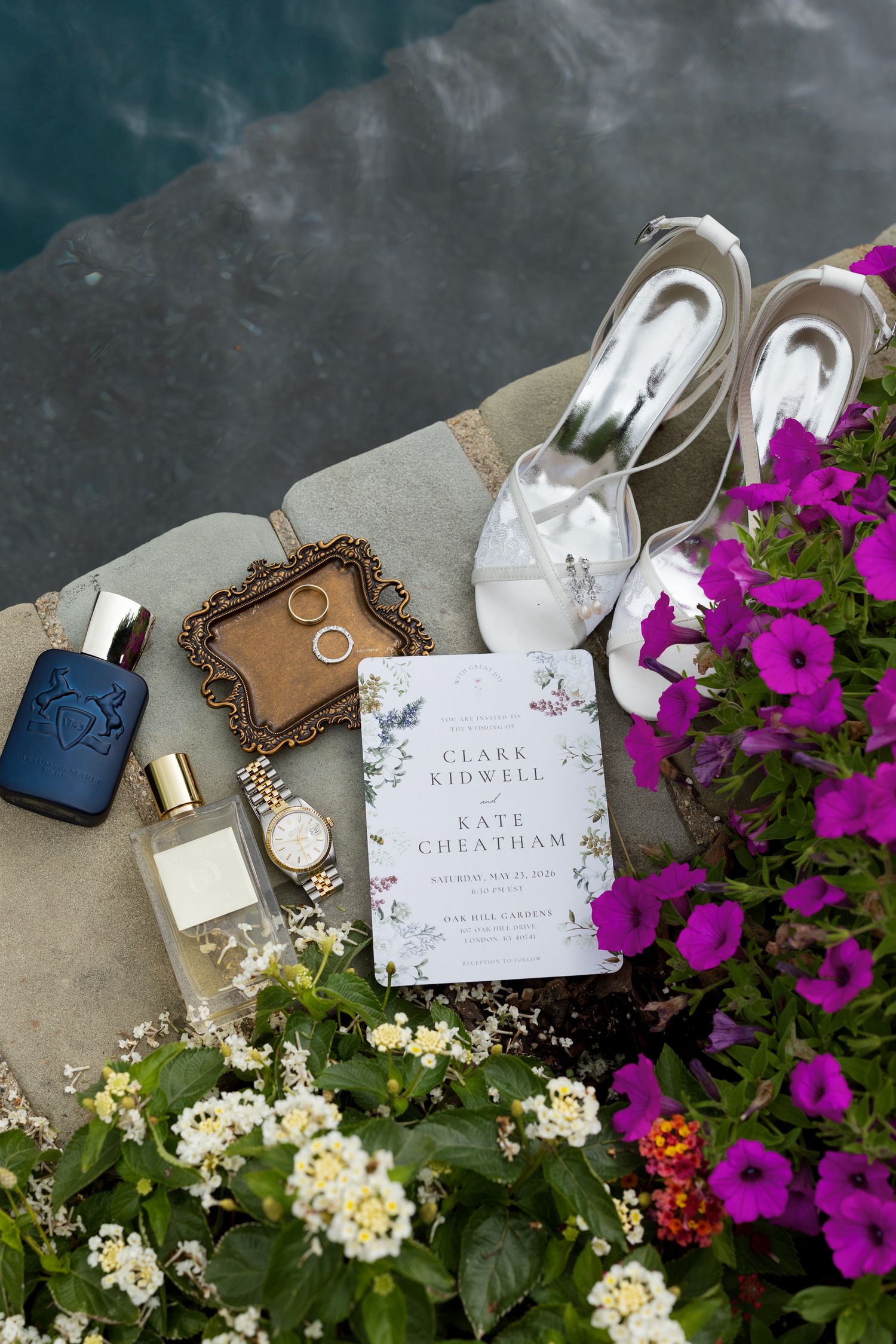 Wedding flat lay featuring invitation for Clark Kidwell and Kate Cheatham, white heels, rings, perfume, watch, and purple flowers.