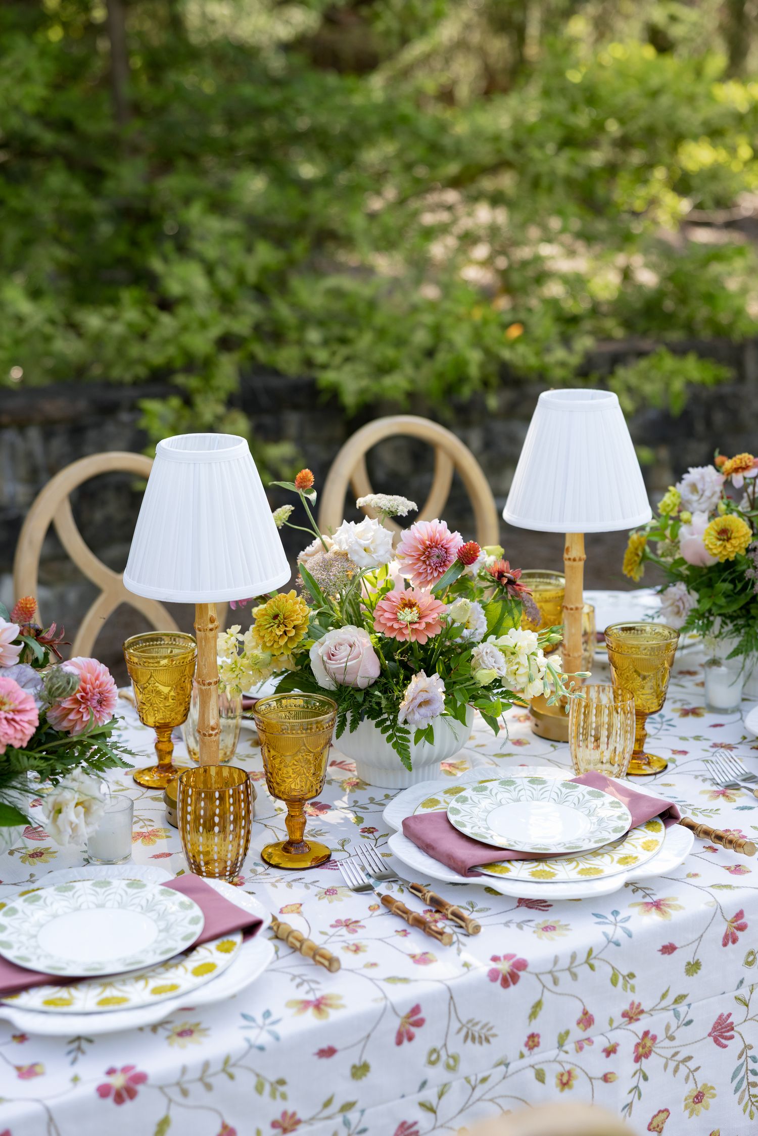 Elegant outdoor tablescape with floral tablecloth, amber glasses, small lamps, and pink flower centerpiece.