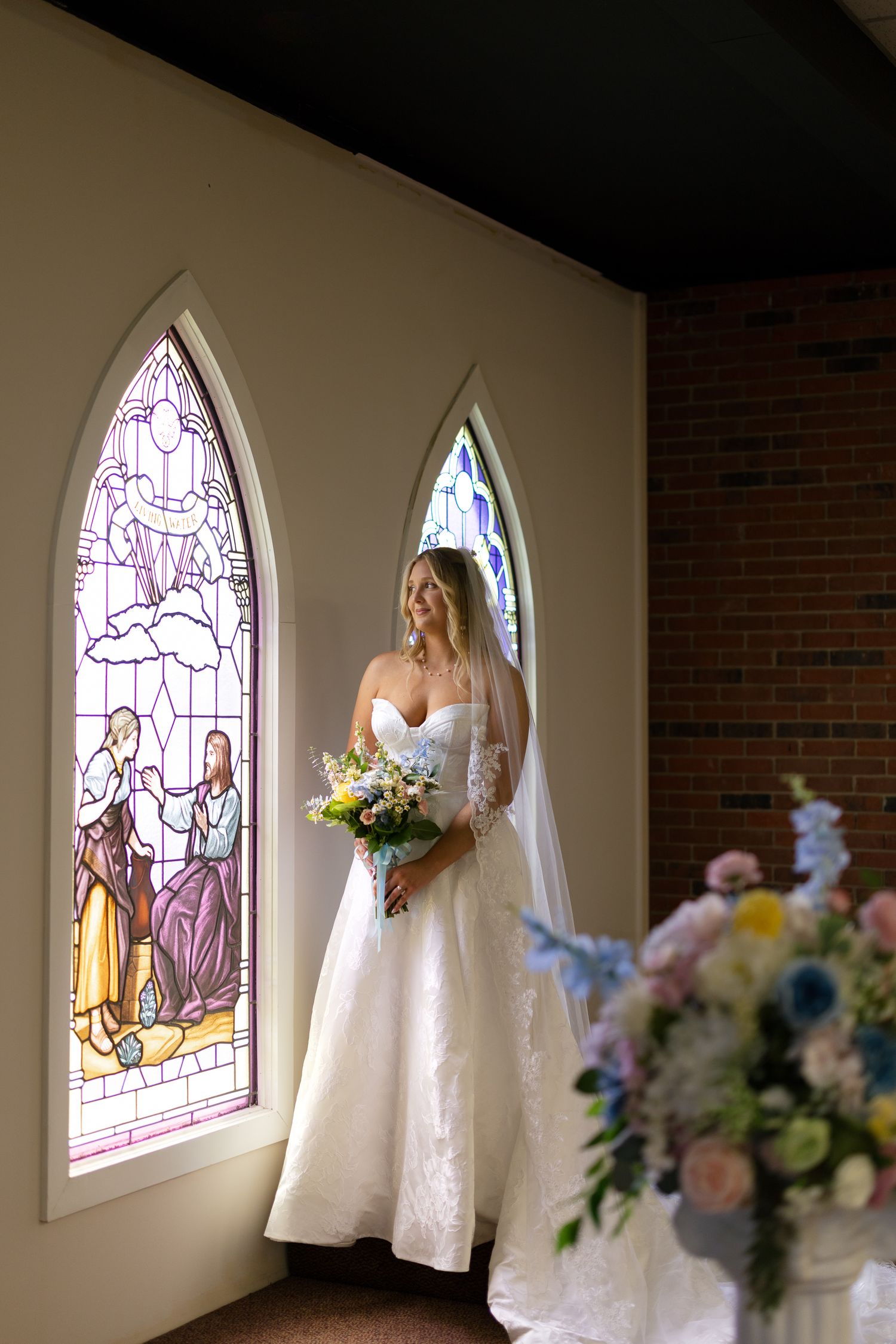 Bride in white gown and veil holds bouquet by stained glass windows in a church.