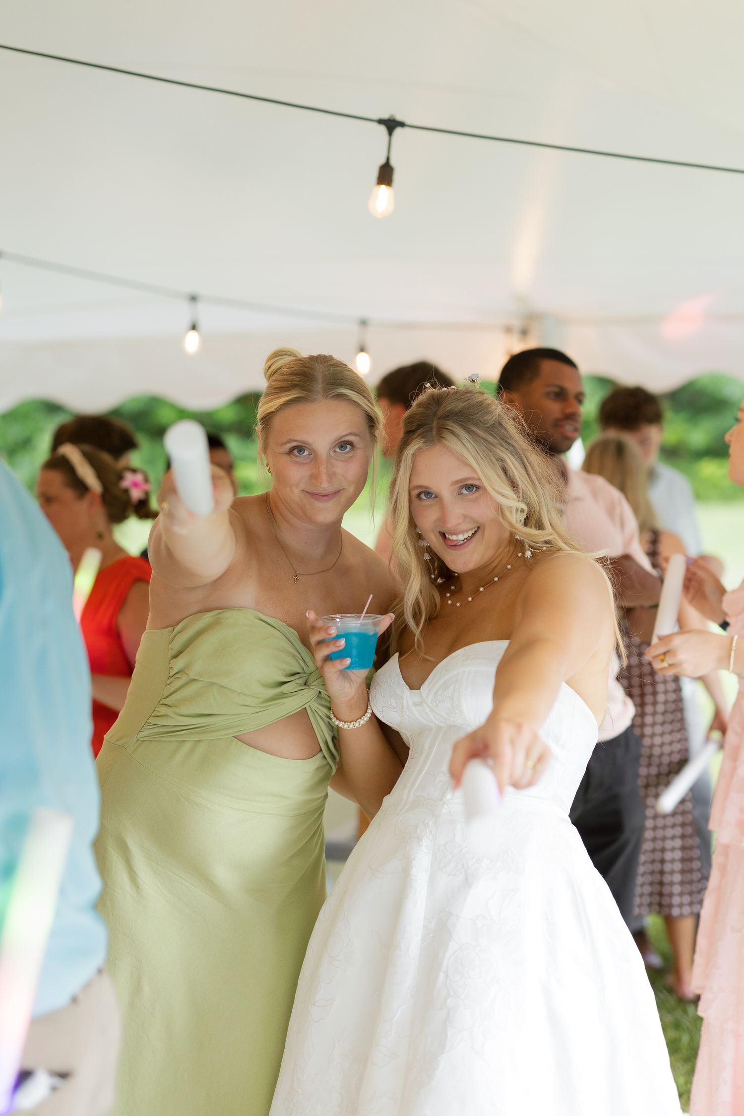 Bride in white gown and bridesmaid in green dress pose and point at camera during outdoor wedding reception.