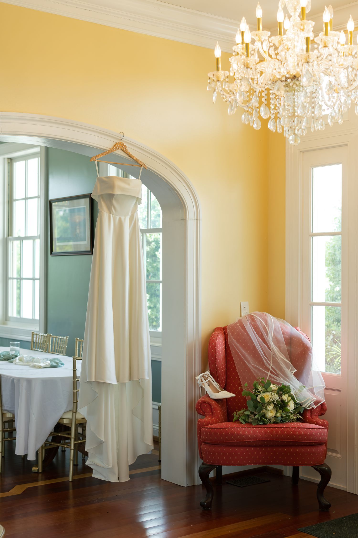 Elegant bridal suite with white wedding dress hanging in arch, red chair with veil and bouquet, crystal chandelier.