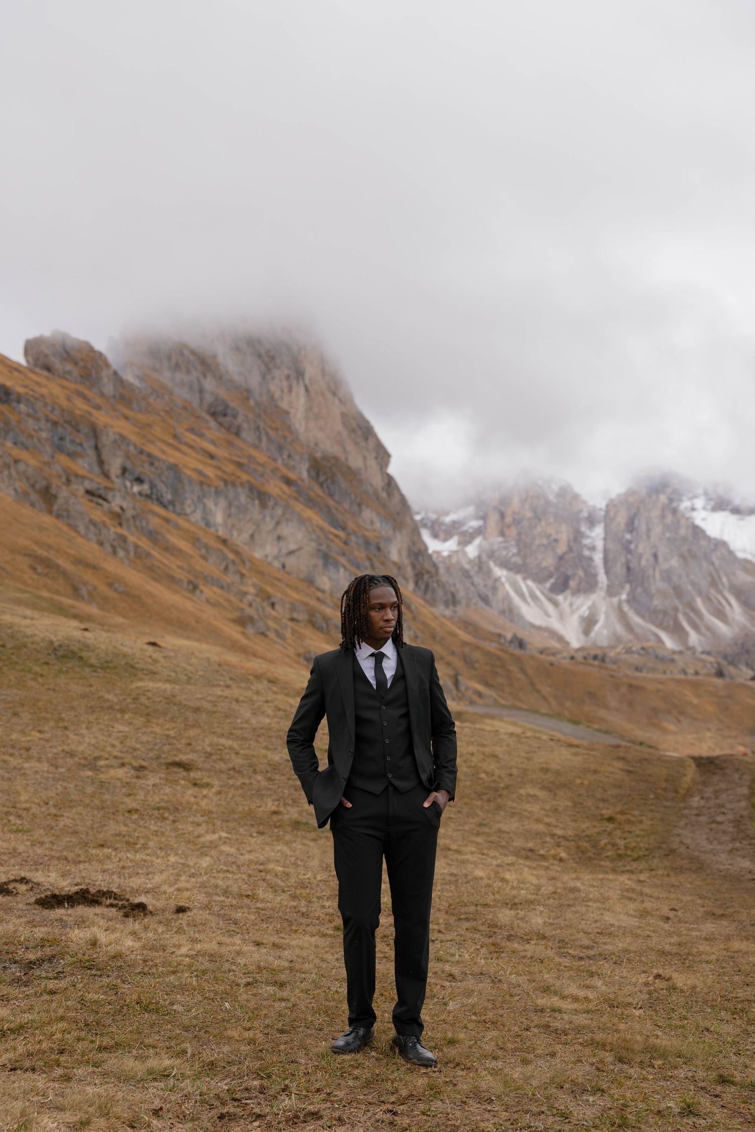 A groom in a dark suit stands on a mountainside with misty peaks in the background in the Dolomites at Seceda