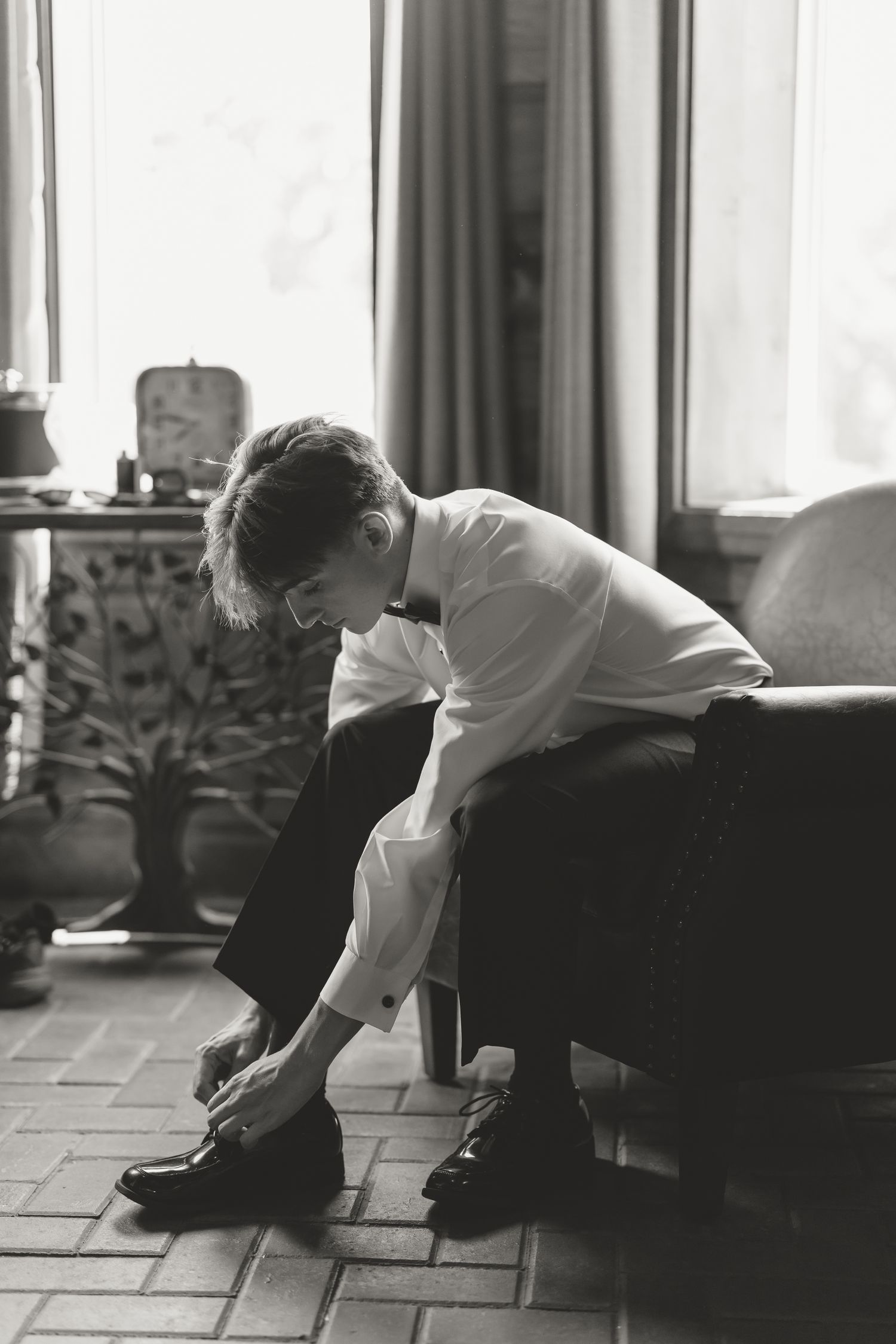 Black and white photo of a young man in formal wear sitting on a chair, tying his dress shoes in an elegant room.