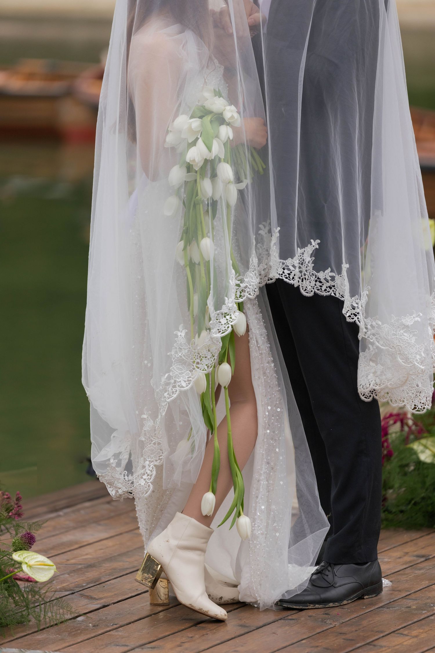 Close-up of bride and groom's feet with white lace veil and calla lily bouquet on wooden deck.