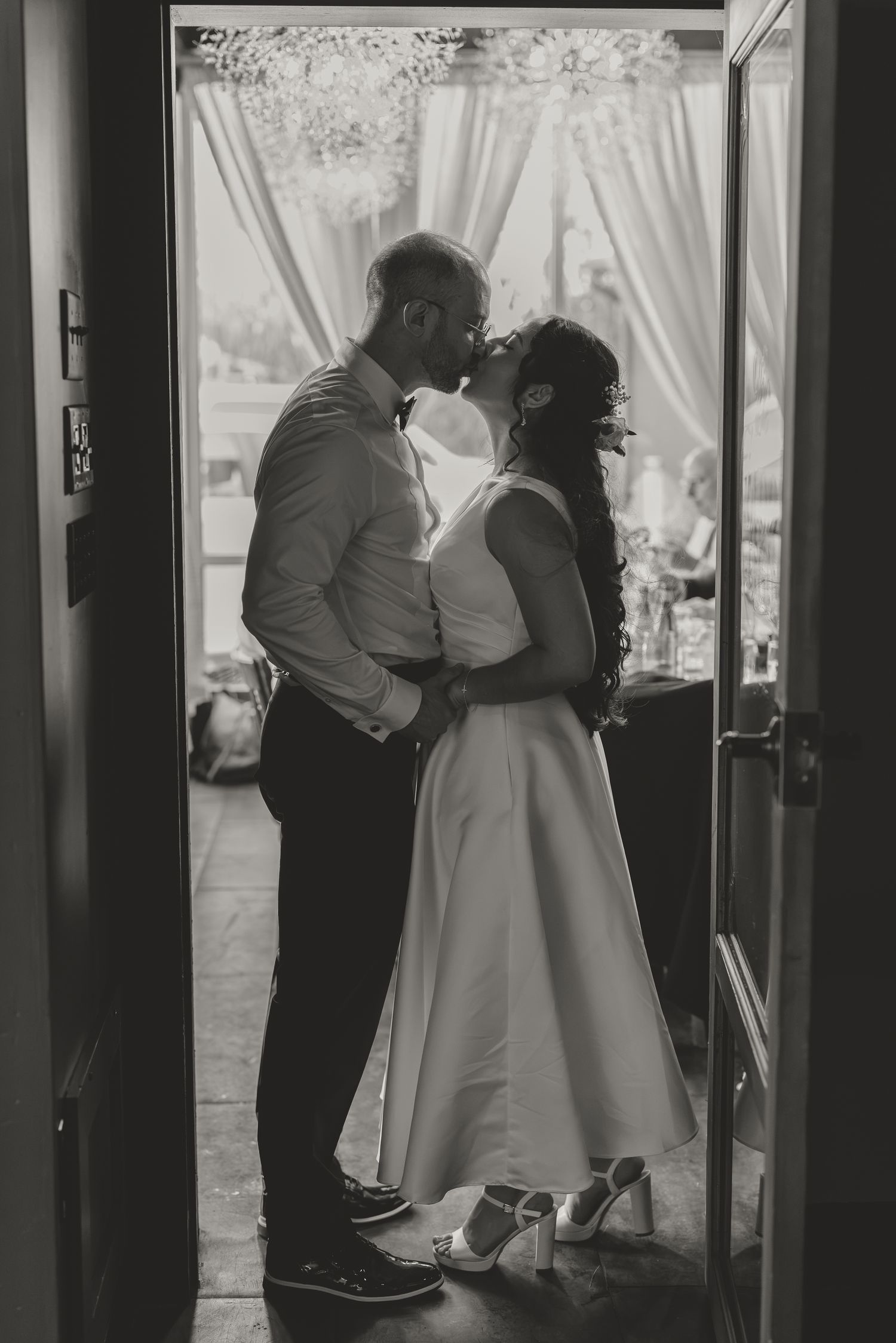 Black and white photo of a couple sharing a romantic kiss in a doorway, bride in white dress and groom in suit.